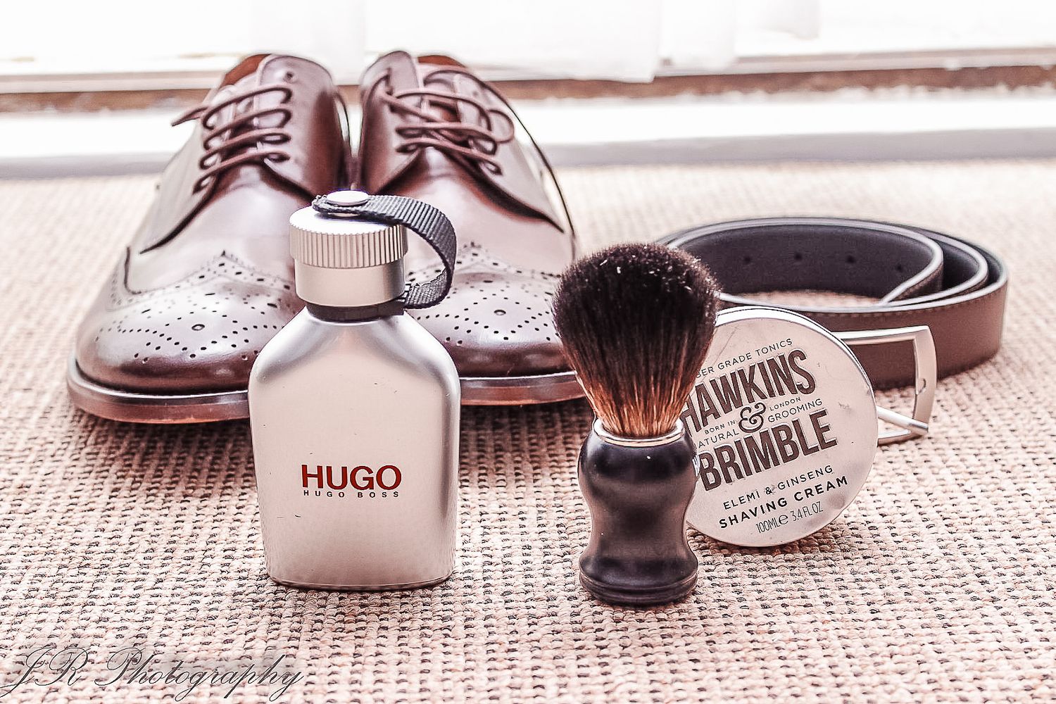 Wedding accessories including brown leather shoes, shaving brush, cologne bottle and belt arranged on fabric.