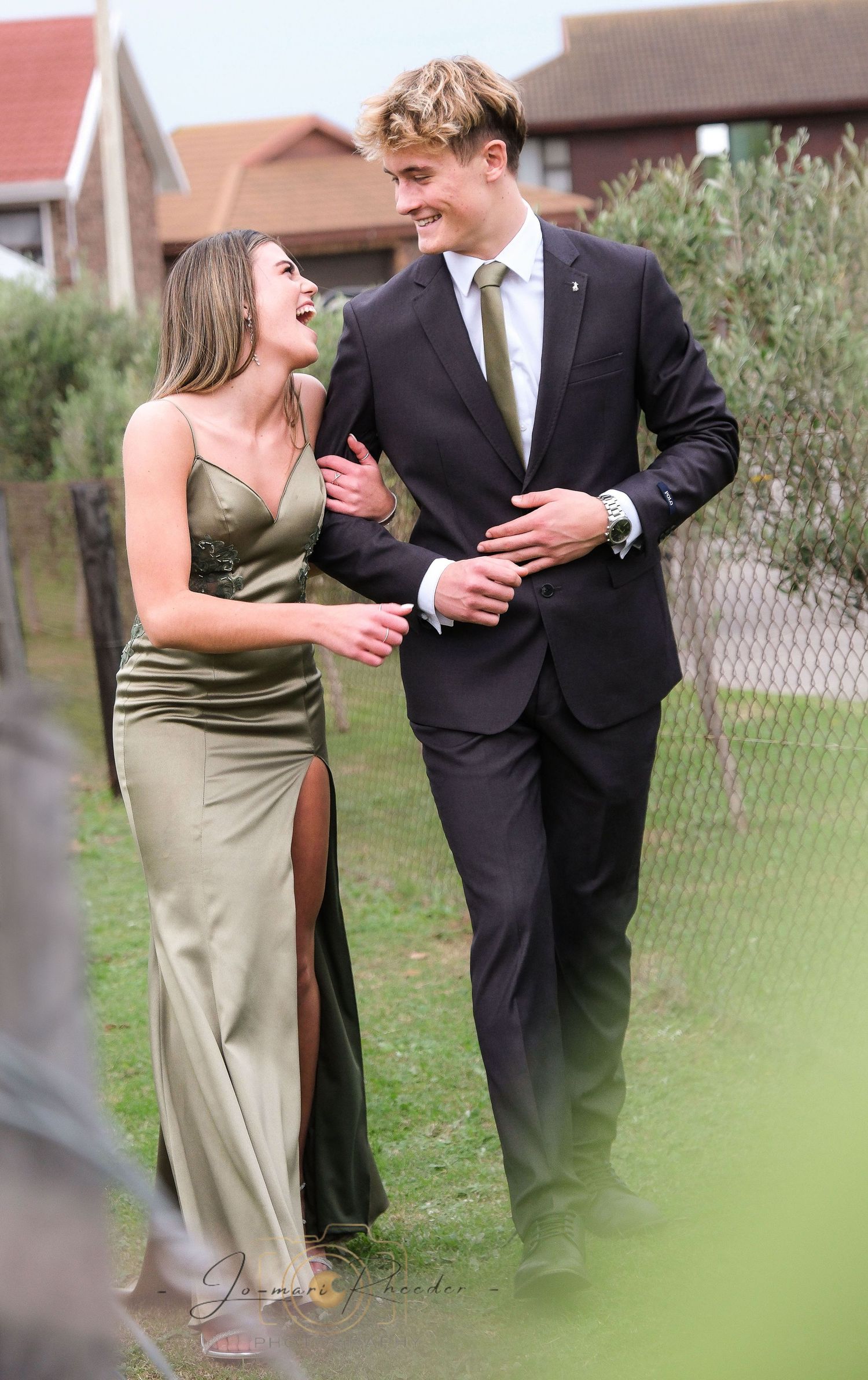 A couple shares a laugh while walking together, dressed formally in olive green and black.