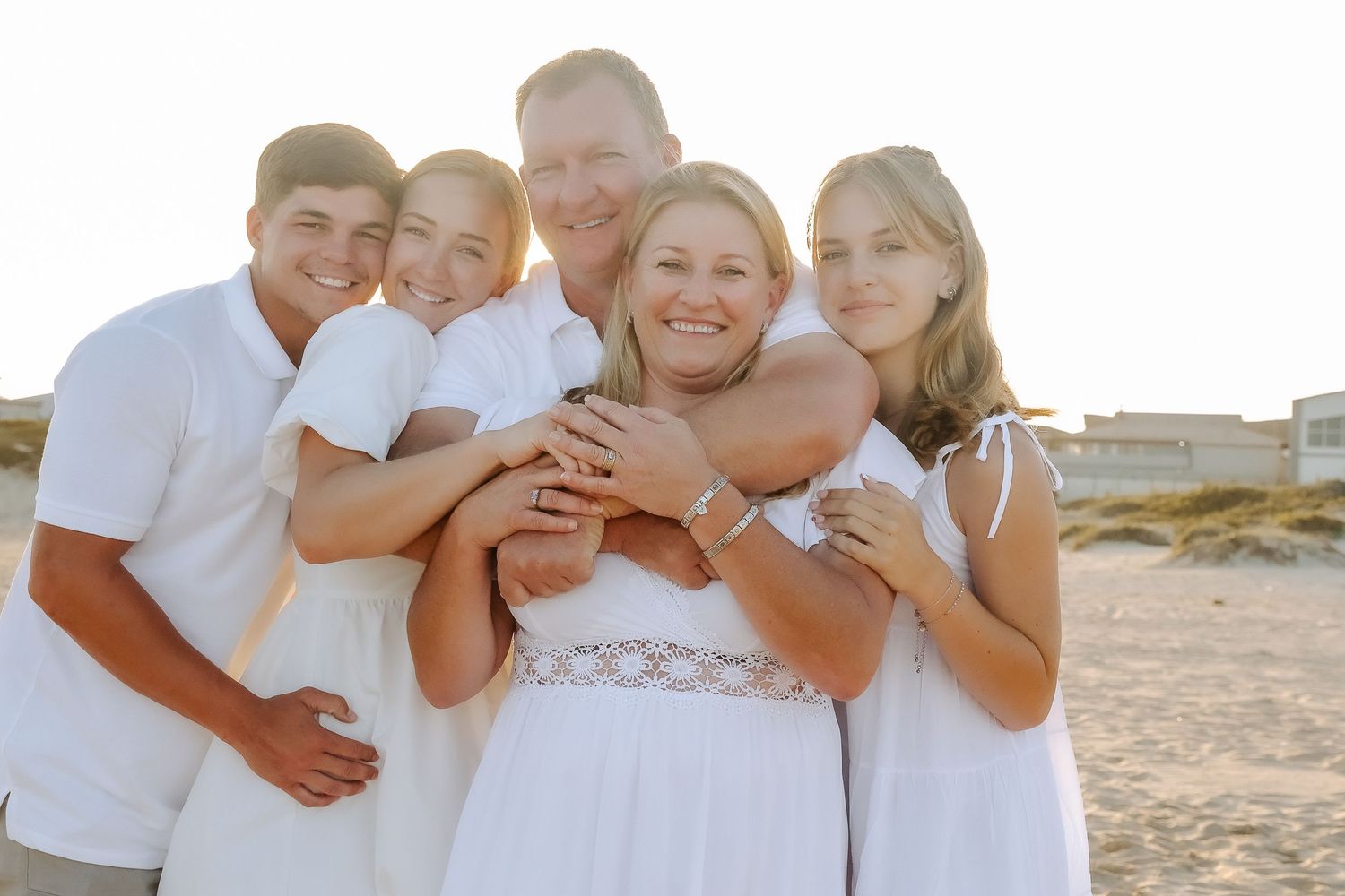 A family group embraces in white outfits during a beach photoshoot at golden hour.
