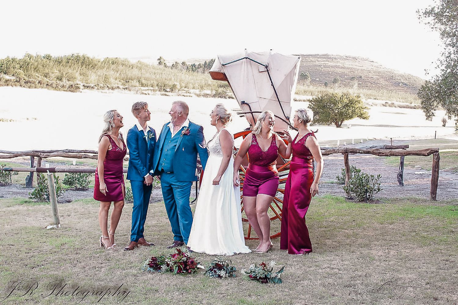 A wedding party poses outdoors wearing burgundy and blue attire against a rural backdrop.