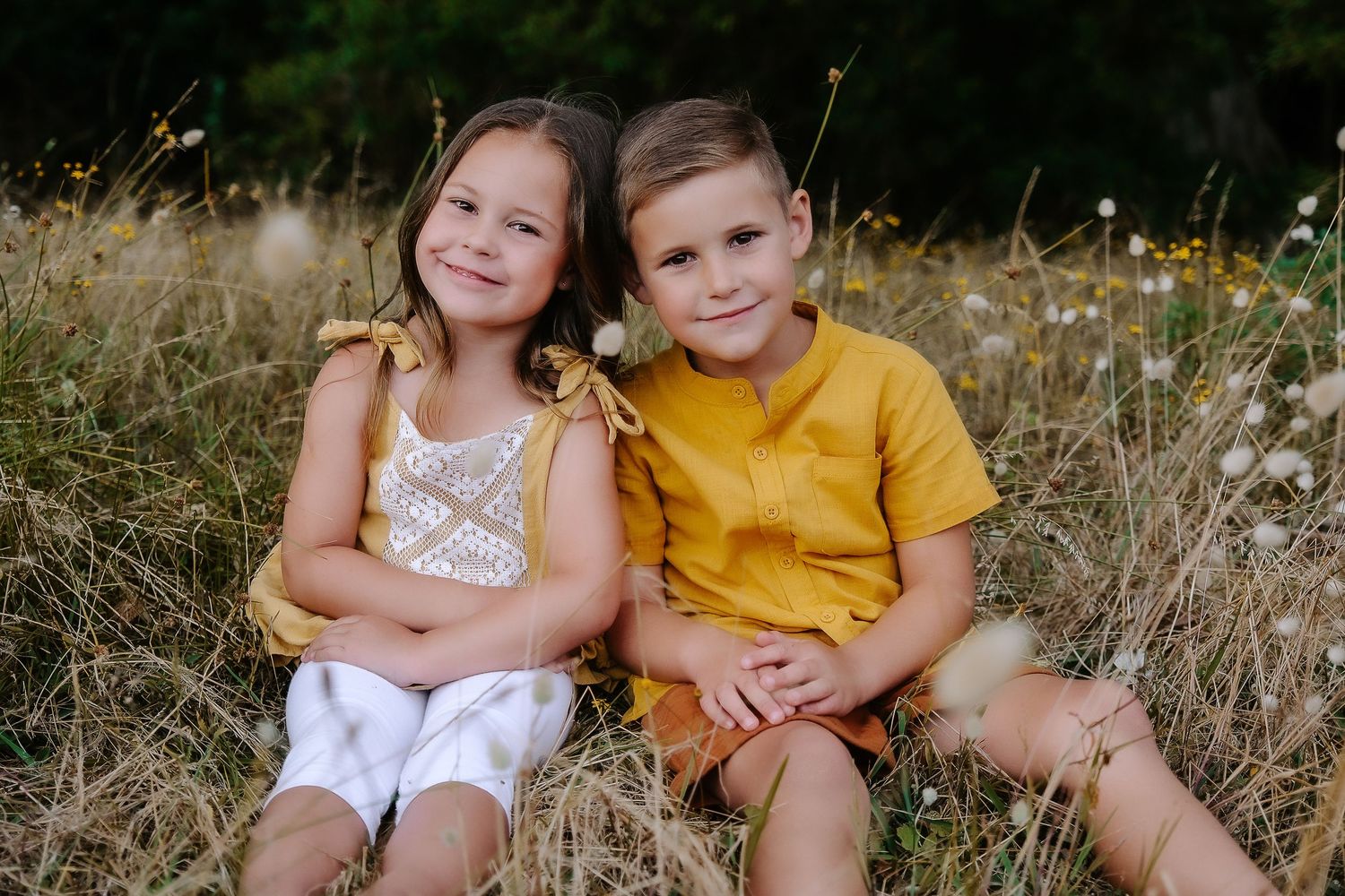 Two young children sitting together in a sunny field with wild grasses.
