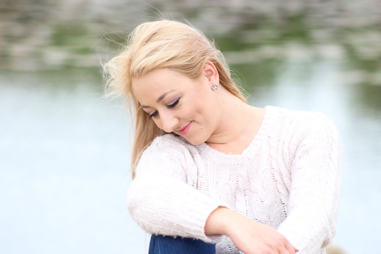Person in white sweater and blue dress resting outdoors by water.
