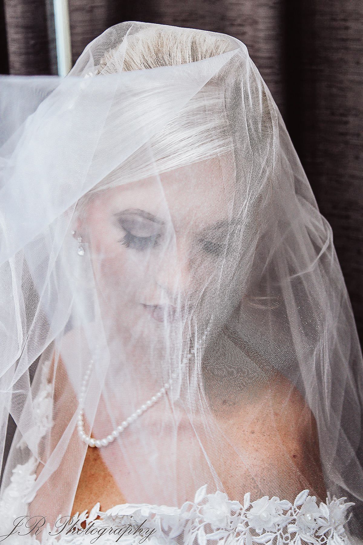 Close-up of bride wearing delicate white veil with soft makeup and pearl necklace.