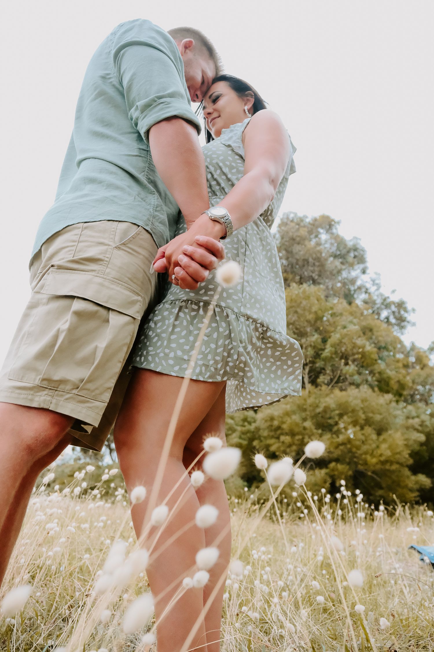 Intimate couple embrace while standing in summer field with tall grass and flowers.