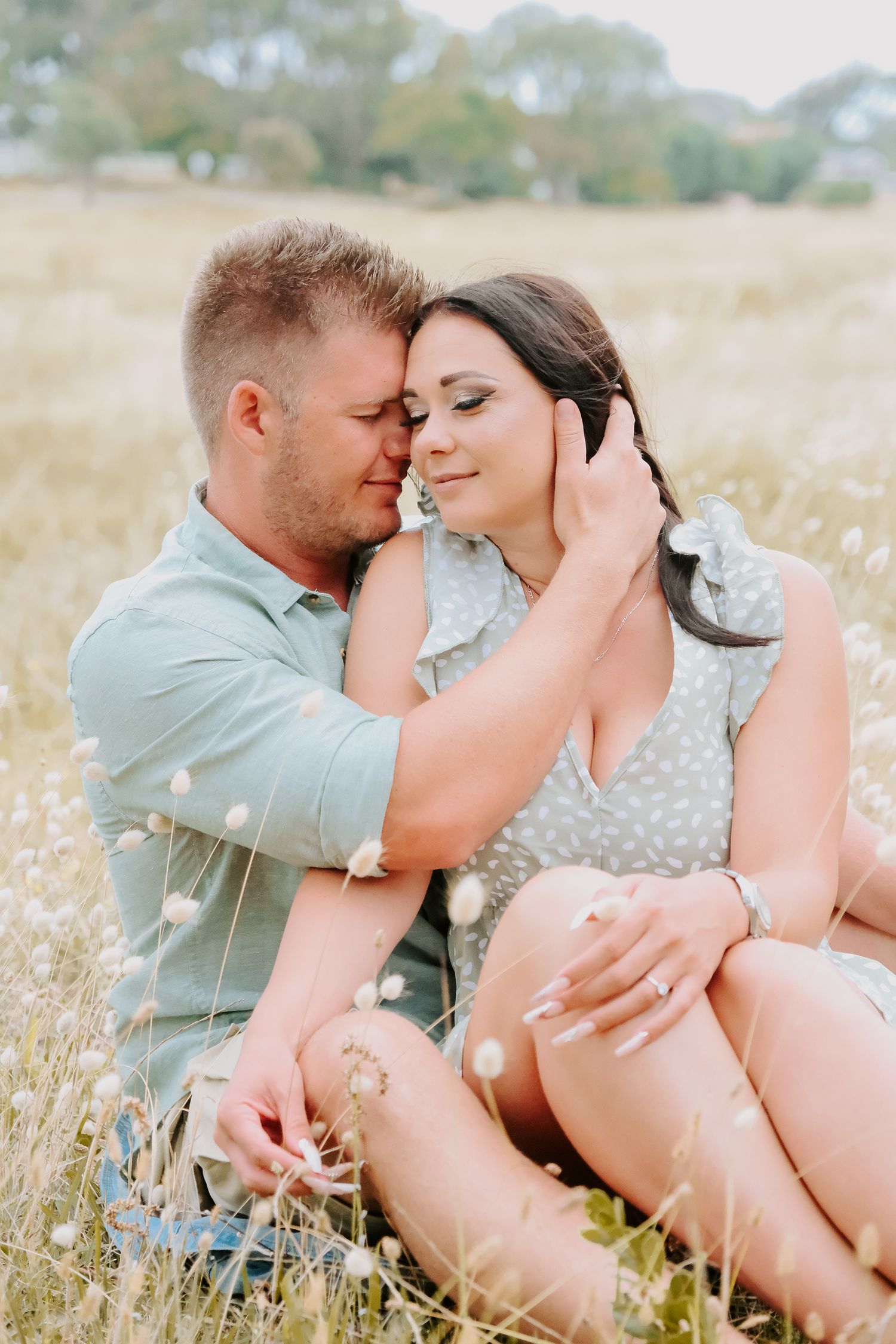 A romantic couple sits together in a sun-drenched meadow filled with golden grass during a warm afternoon.