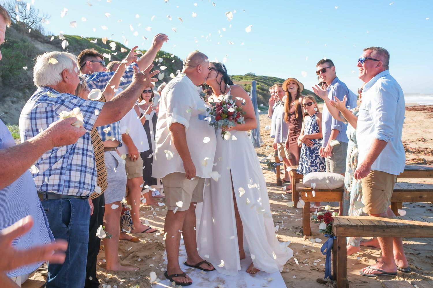 Wedding guests throw confetti over couple during beachside ceremony with bright blue sky and coastal backdrop.