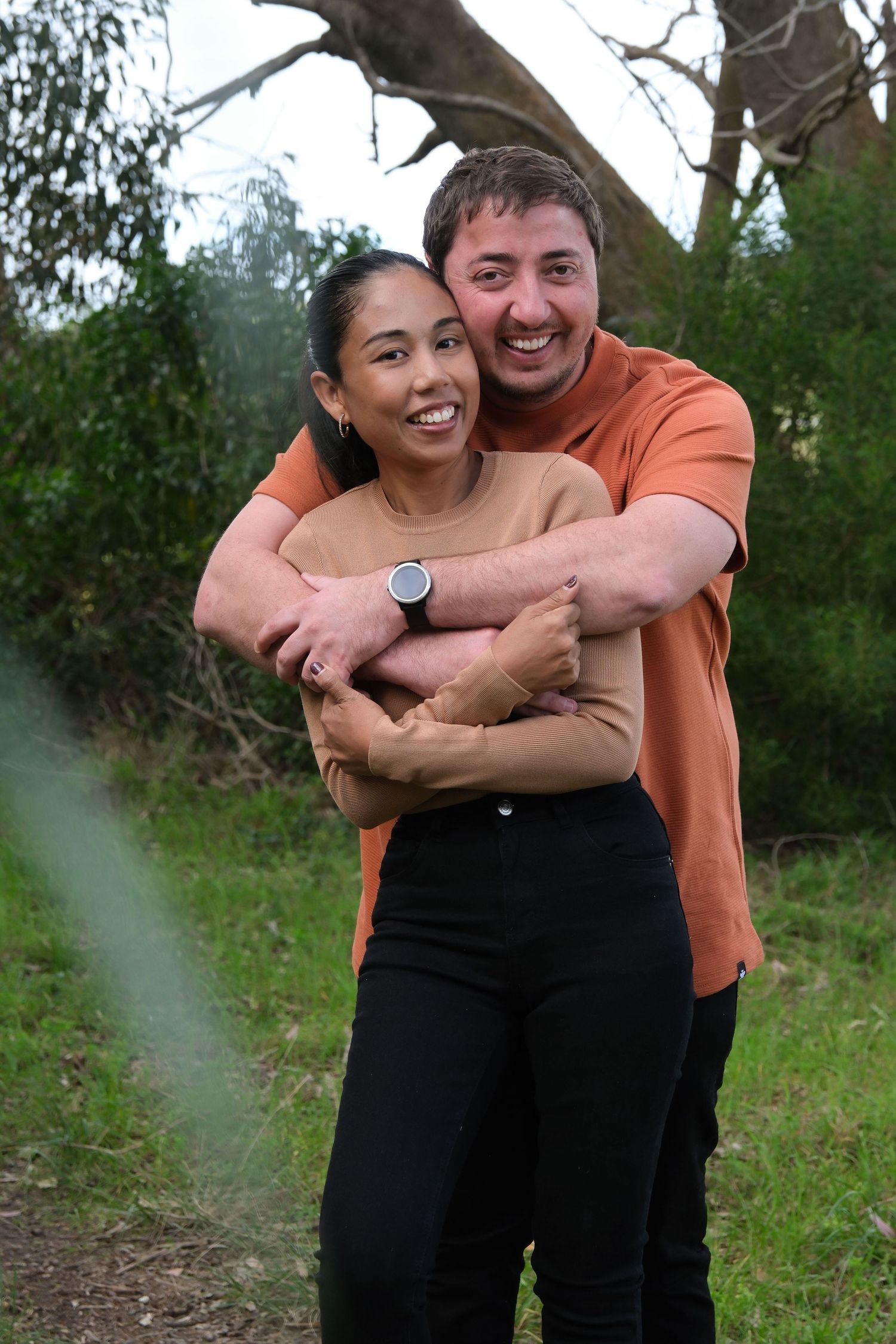 A couple embraces and smiles while standing outdoors in a natural setting with trees in the background.