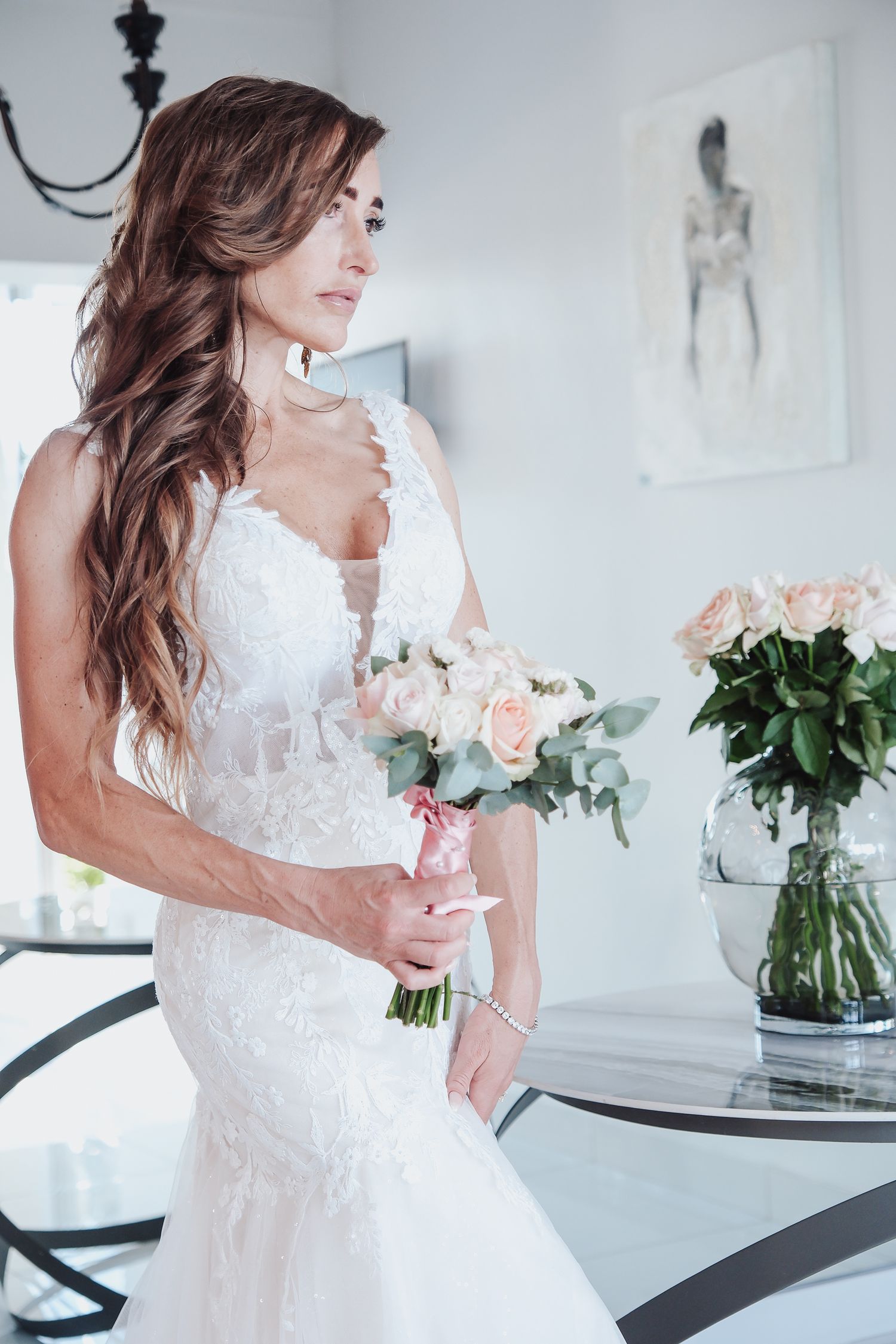 Woman in fitted lace wedding gown holding pink bouquet in elegant white room.