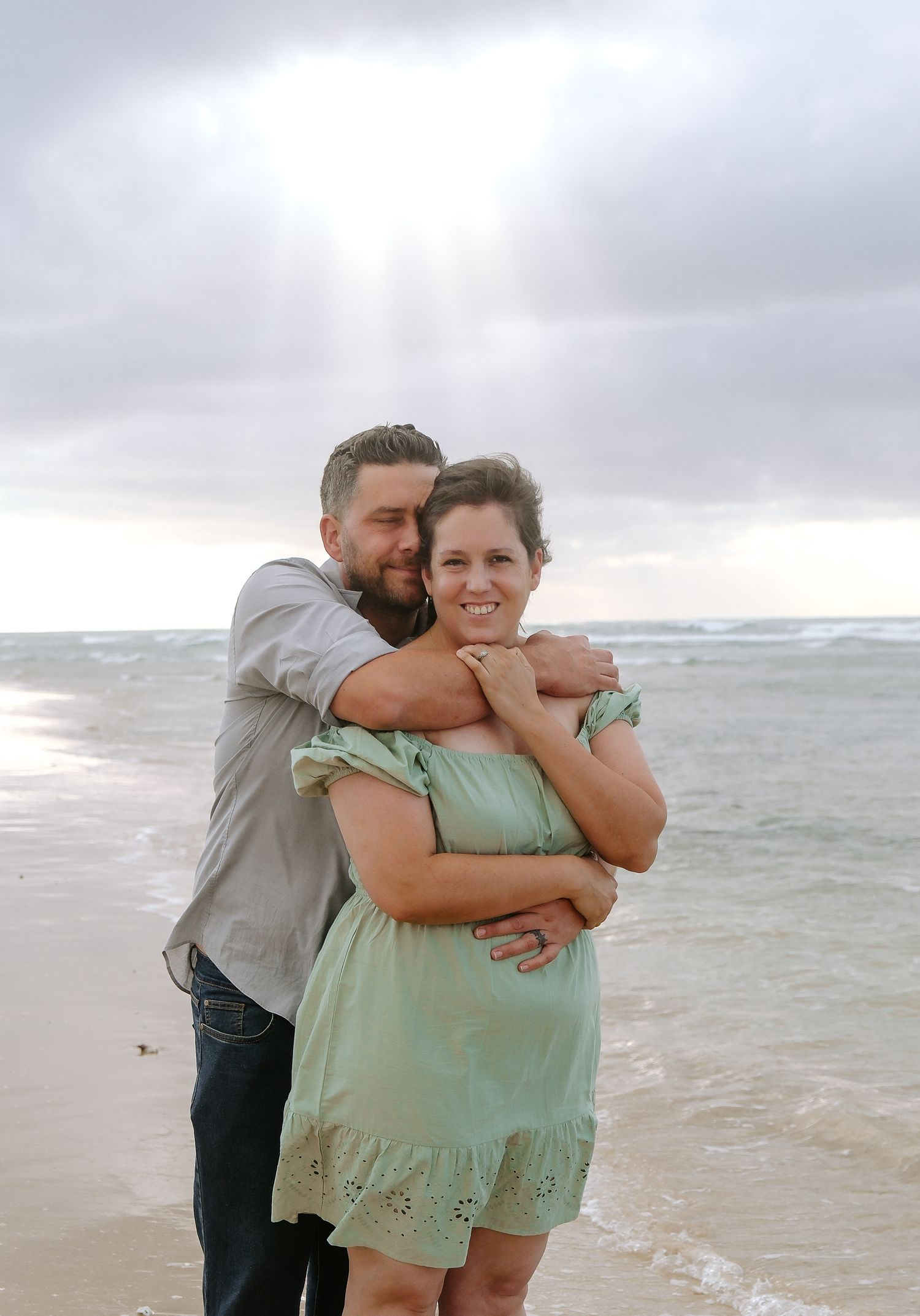 Romantic couple embraces on a cloudy beach with waves in the background.