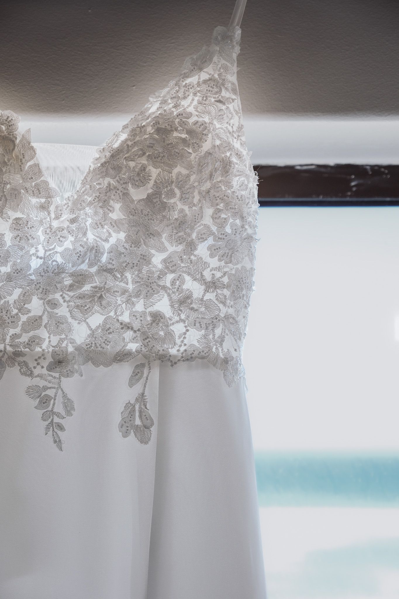 White wedding dress with delicate lace bodice hanging against a light background showing intricate floral detail.