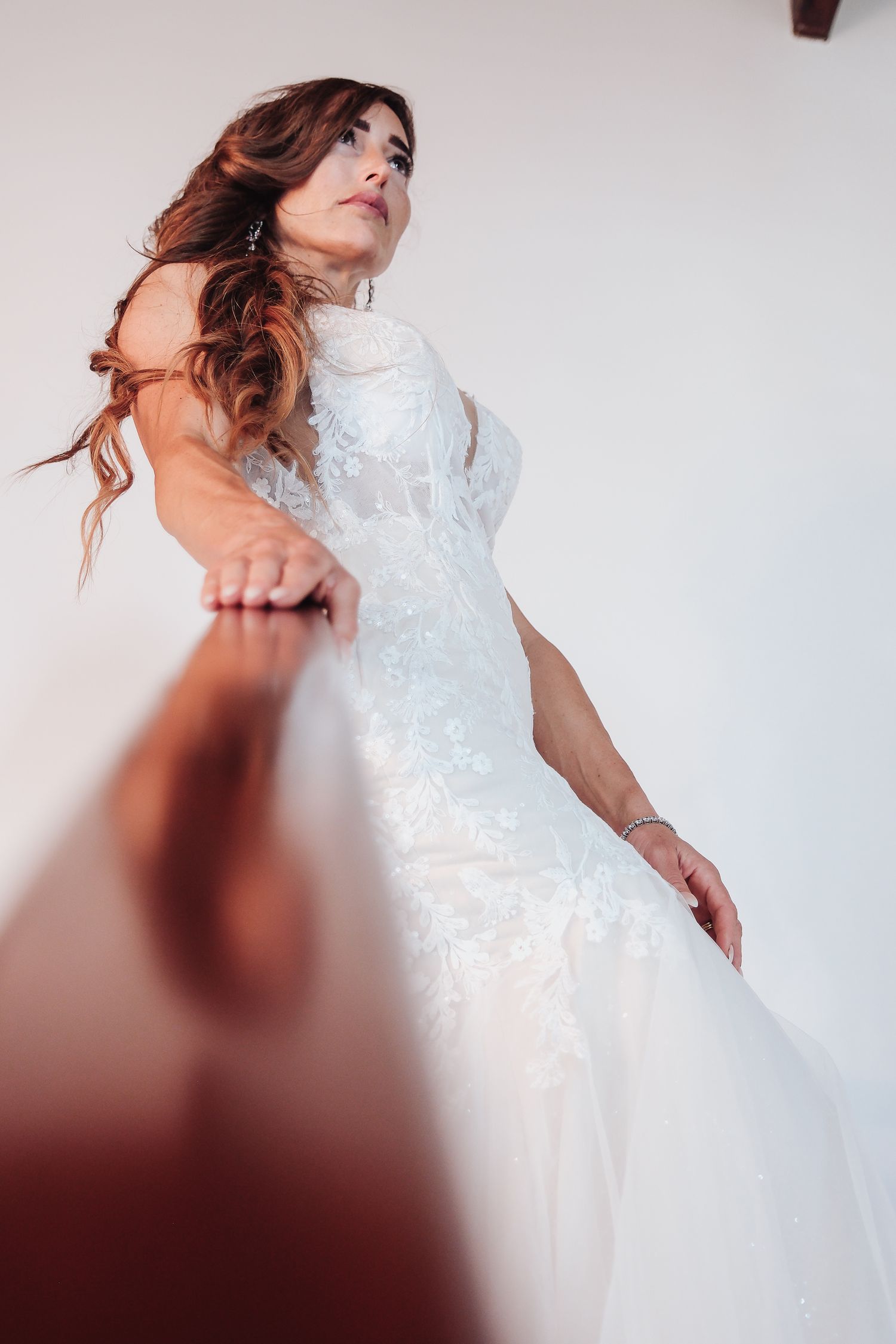 Artistic angle of bride in fitted lace wedding dress against white wall.