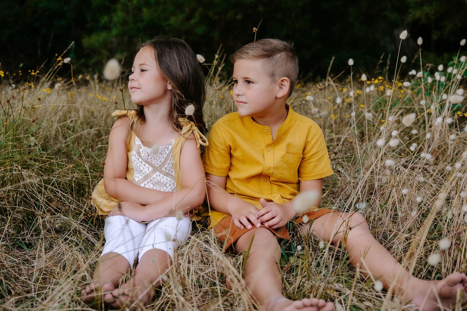 Two children sit back to back in a field of wildflowers wearing summer clothing.