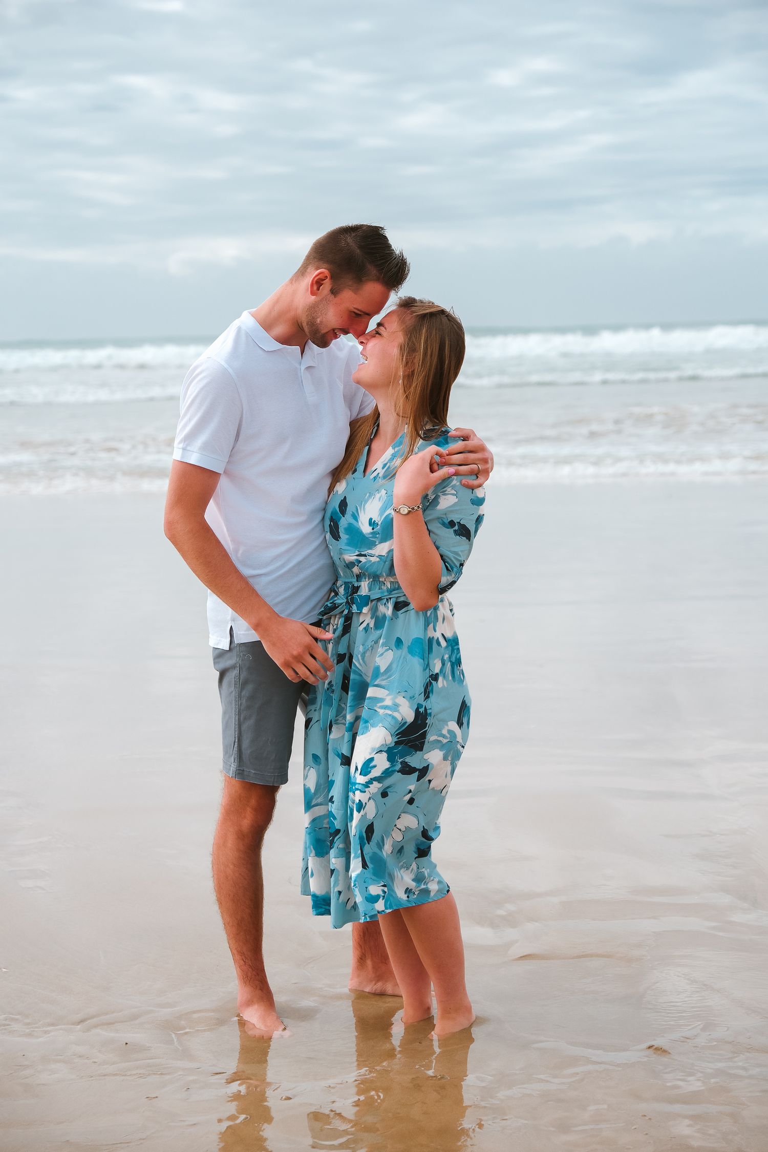 Romantic couple embraces on beach with waves in background, woman wearing blue floral dress.