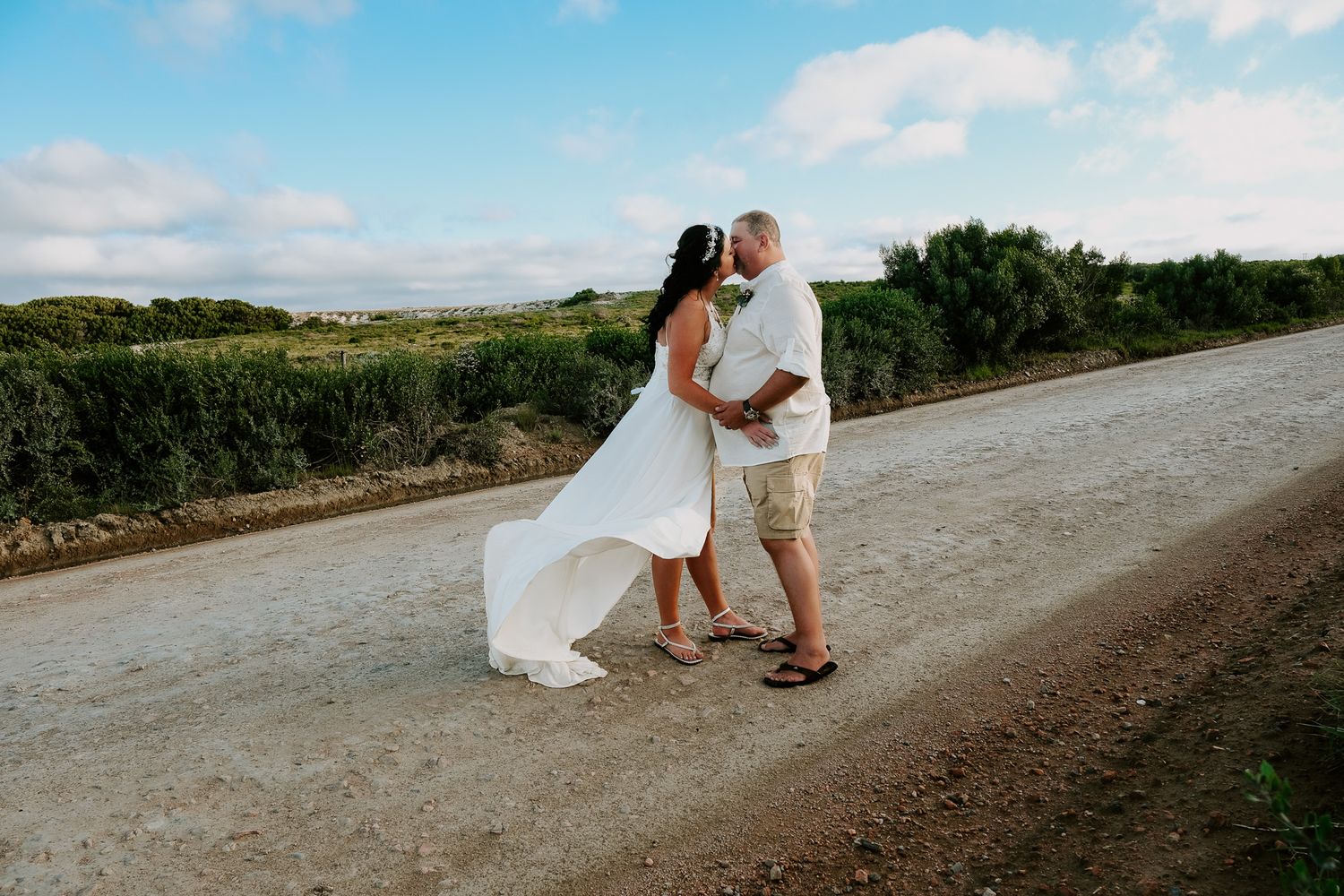 Couple walking together on a dirt path along coastal cliffs surrounded by greenery on a bright sunny day.