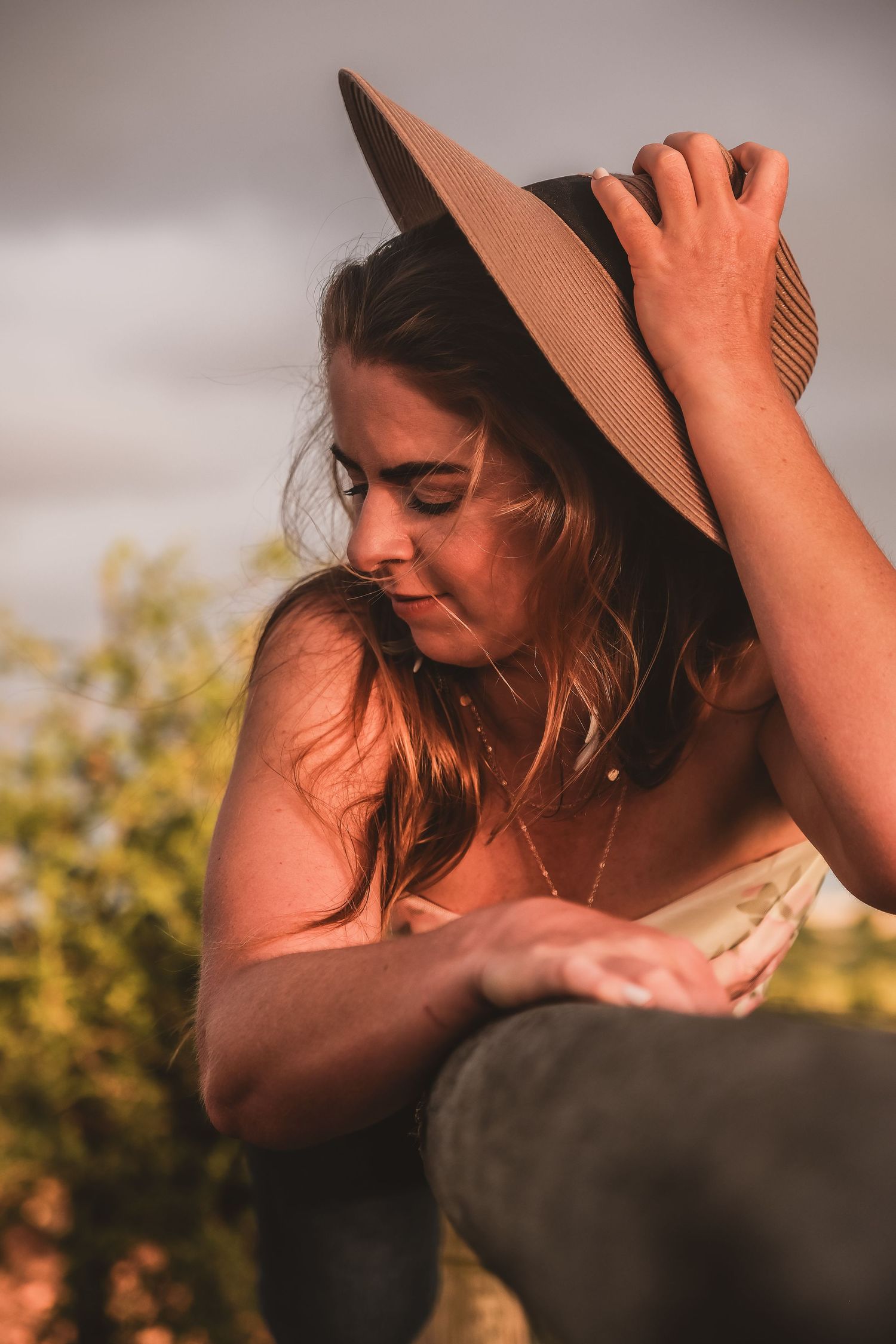 Someone wearing a brown hat smiles while adjusting their hat in golden sunlight.