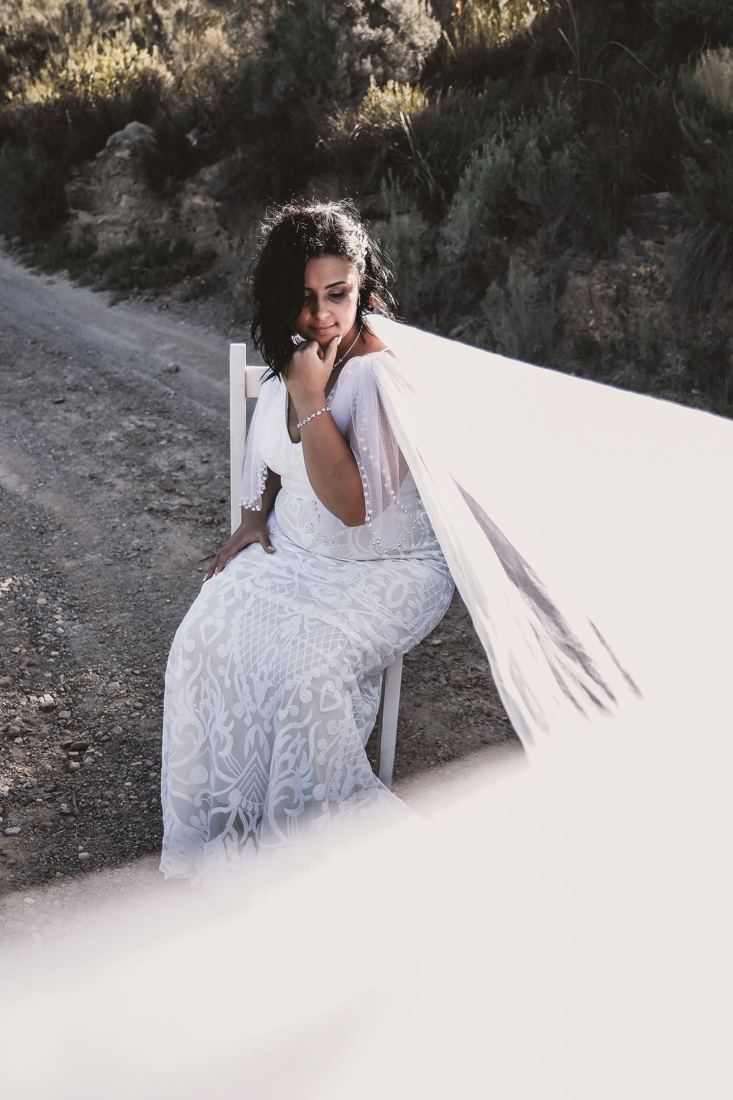 A bride in a detailed lace wedding gown sits thoughtfully on a dirt path against a natural backdrop.