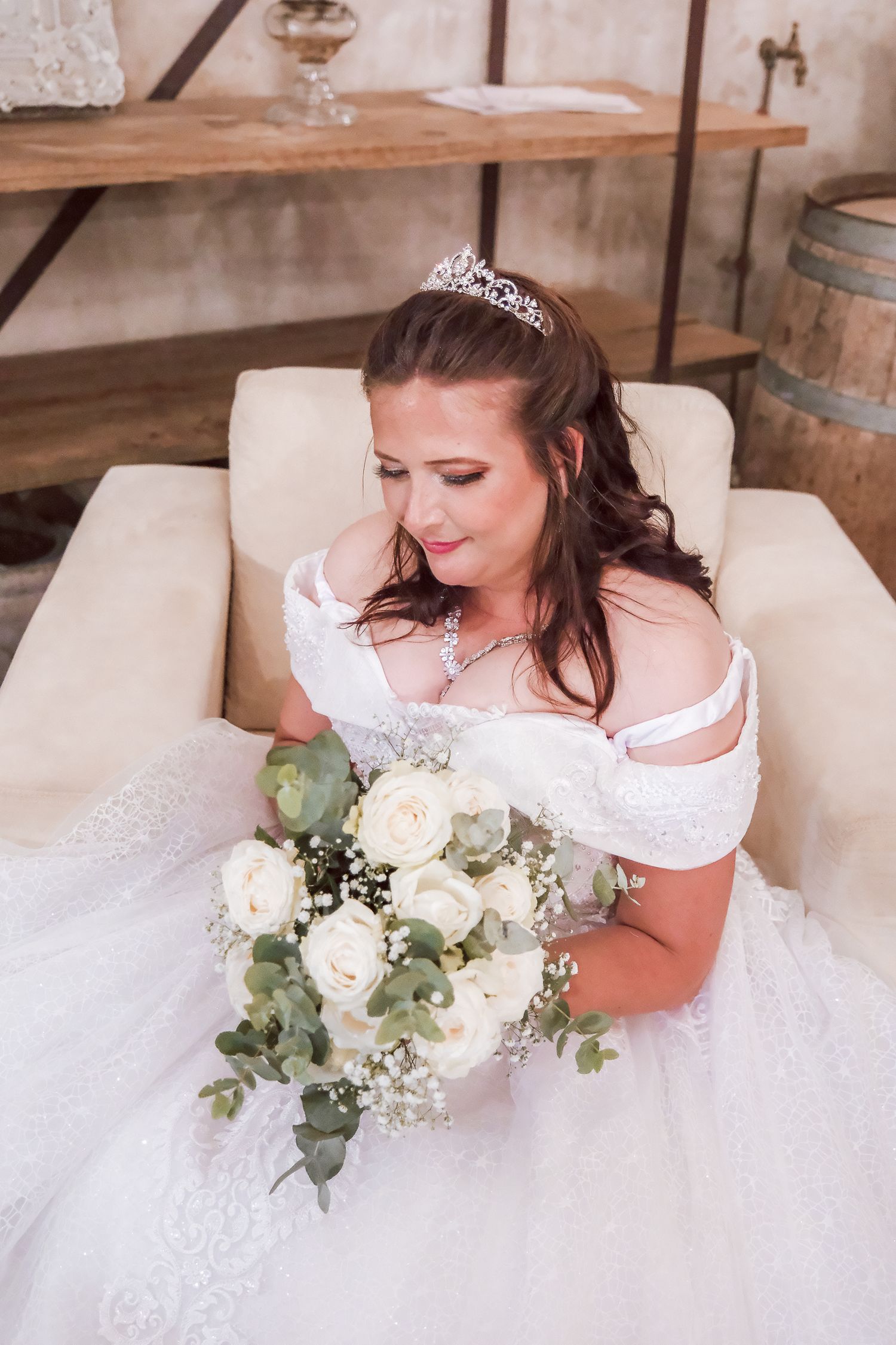A bride in a white gown holds a bouquet of white roses while seated in an elegant chair.