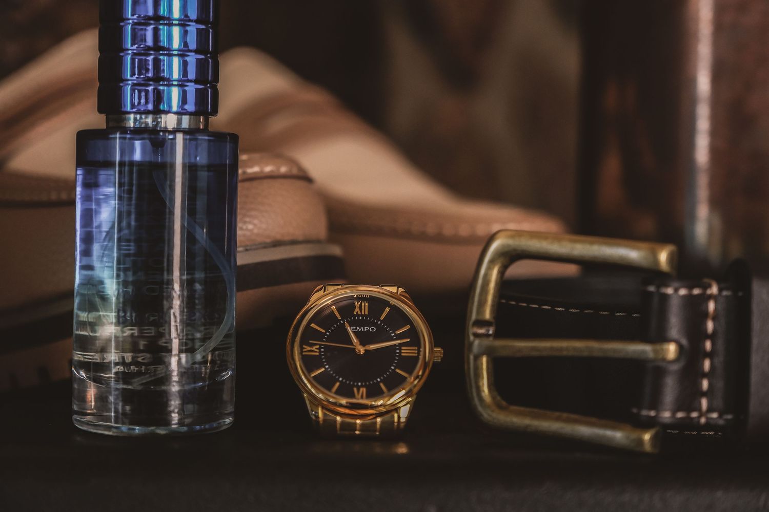 Close-up of luxury watch, cologne, and leather belt arranged on dark surface.