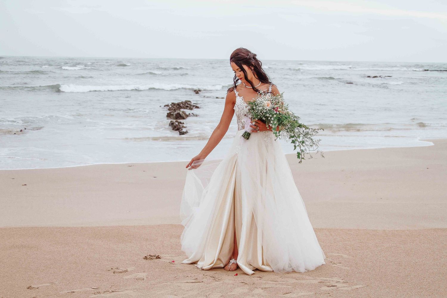 A bride in a flowing white dress holds a bouquet while walking on a windswept beach.