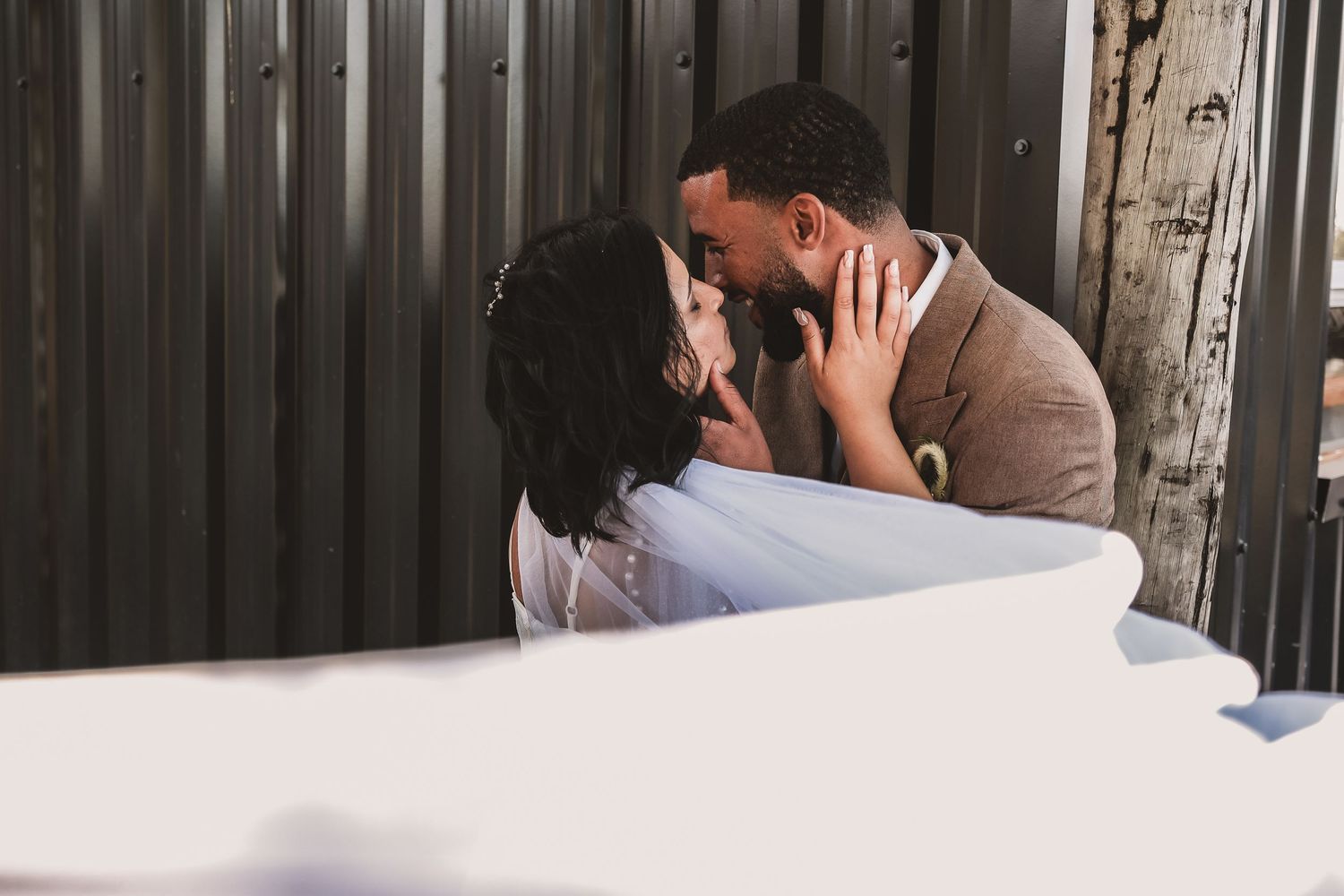 A romantic couple embracing tenderly against a corrugated metal wall in sunlight.