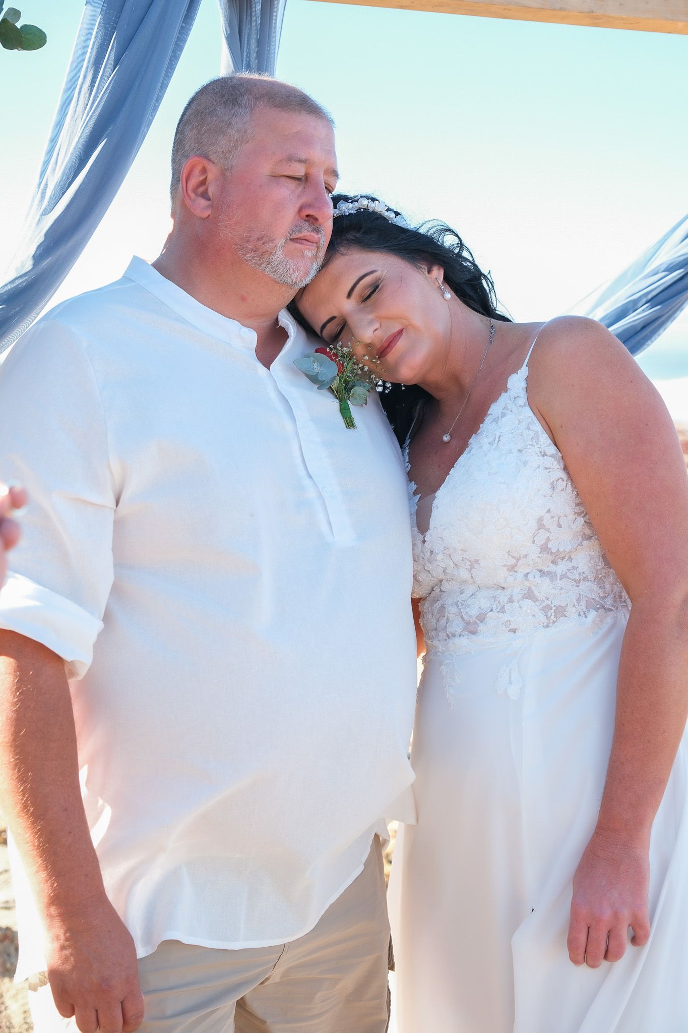 A couple shares an intimate moment under a blue draped canopy, with the person in a lace white wedding dress resting their head tenderly.