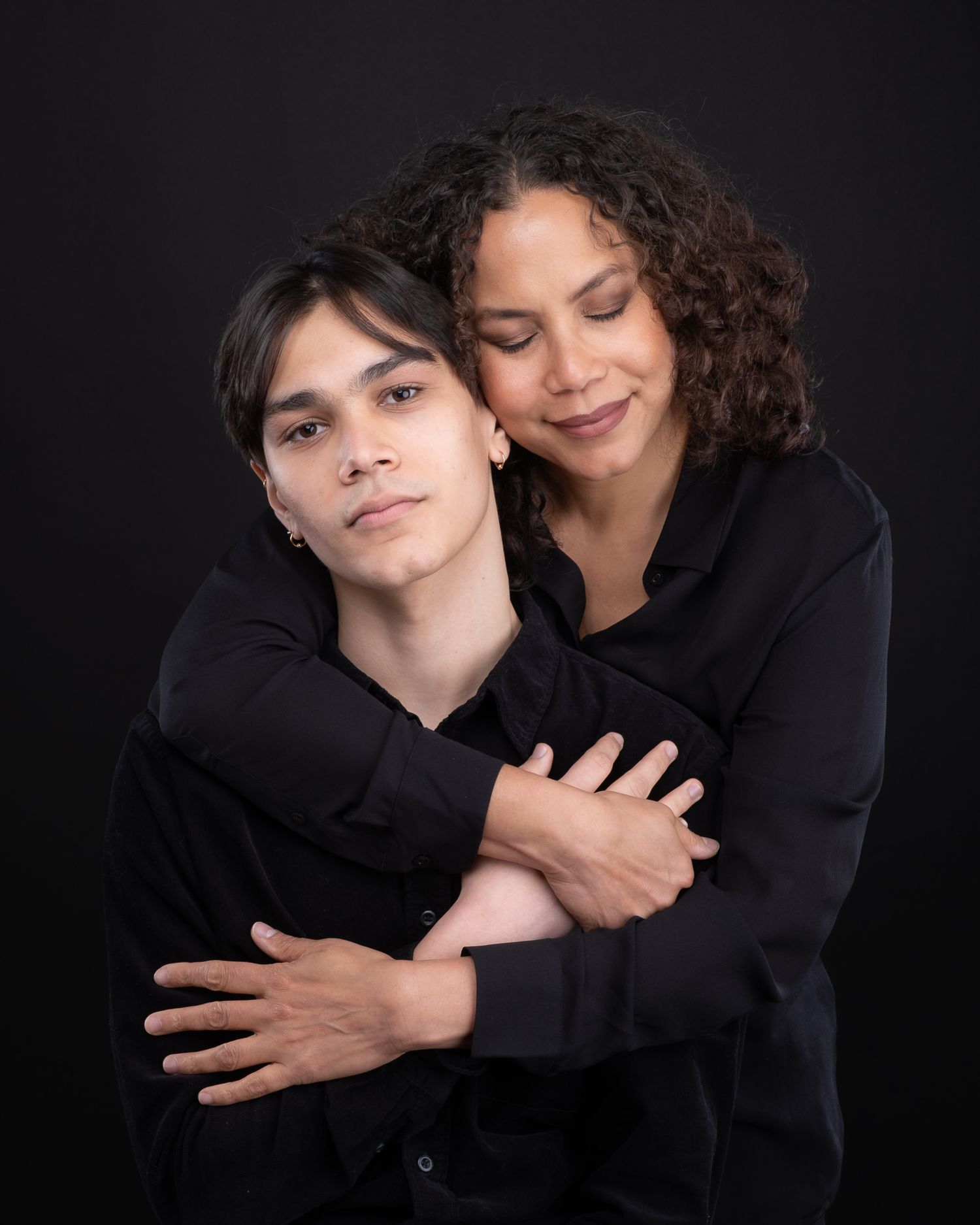 A tender portrait of two people wearing black embracing against a dark background.