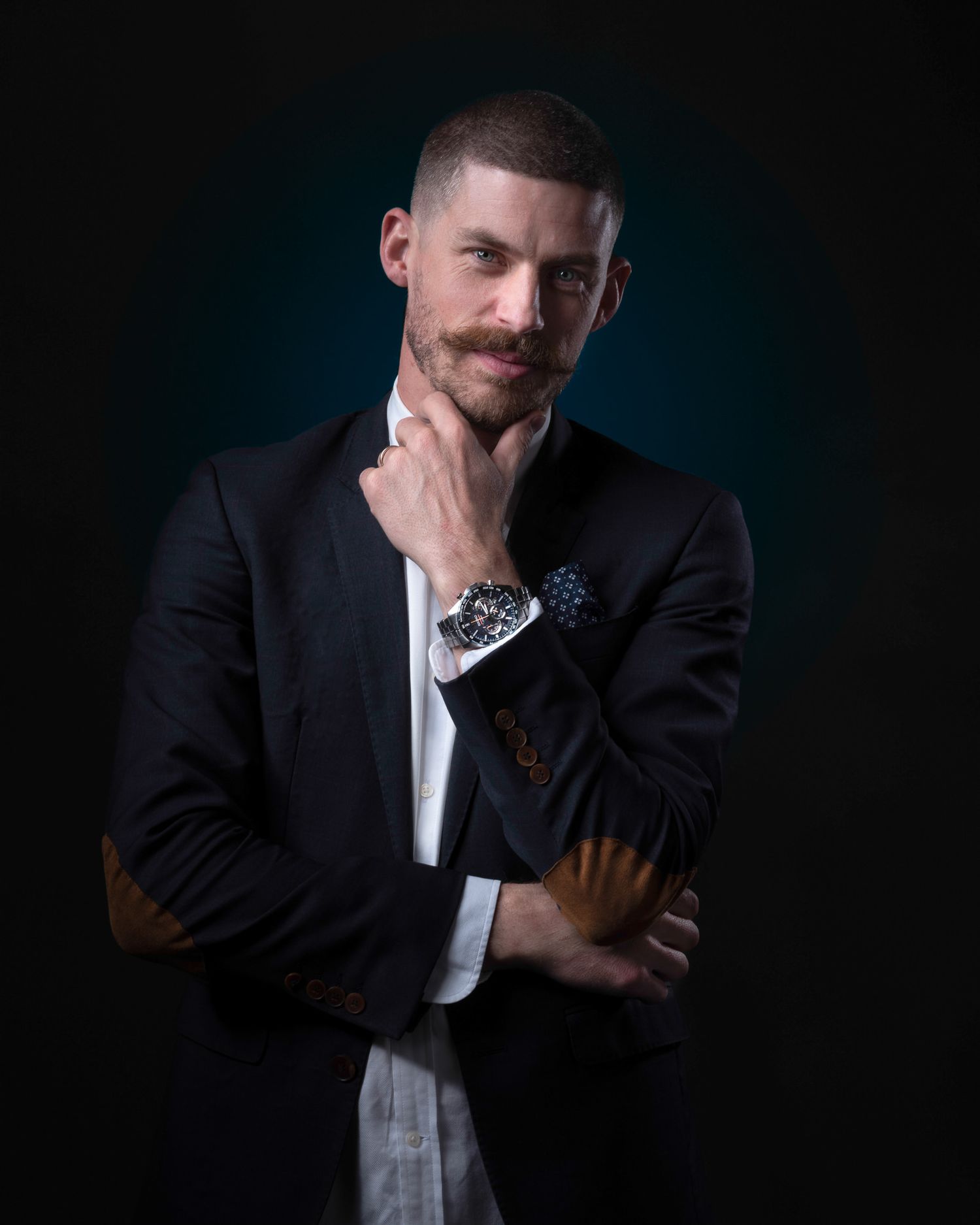Studio portrait of a person in a black blazer and white shirt posing thoughtfully against dark background.