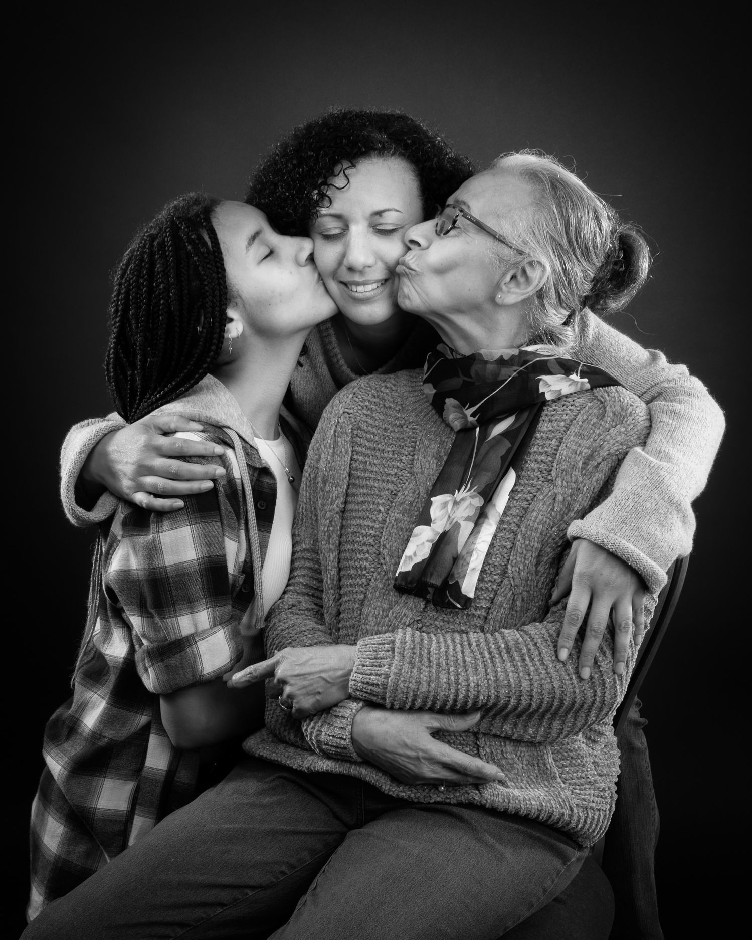 A heartwarming black and white portrait showing three generations sharing loving embraces and kisses in a warm family moment.