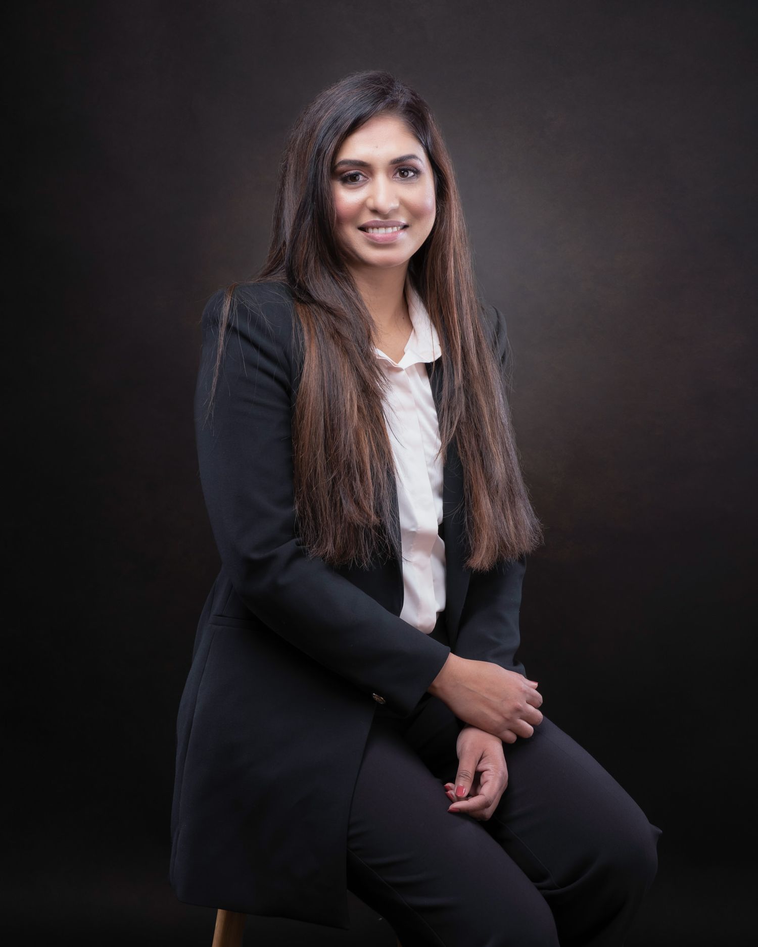 Professional portrait of someone wearing a black blazer and white blouse while seated against a dark background.