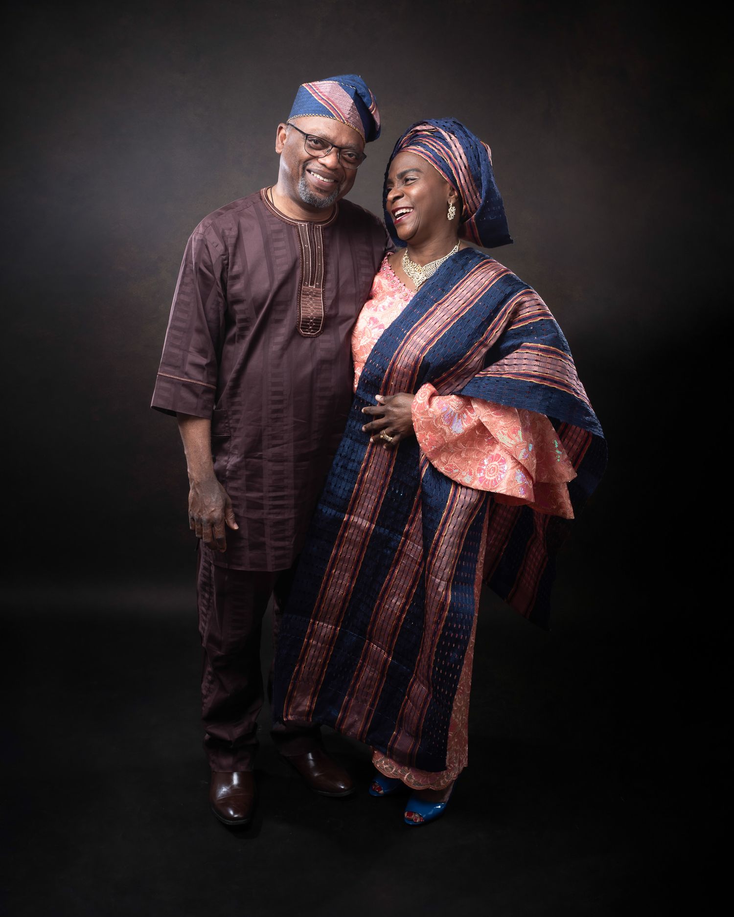 A couple wearing traditional Nigerian attire with matching headpieces pose together against a dark background in a portrait.