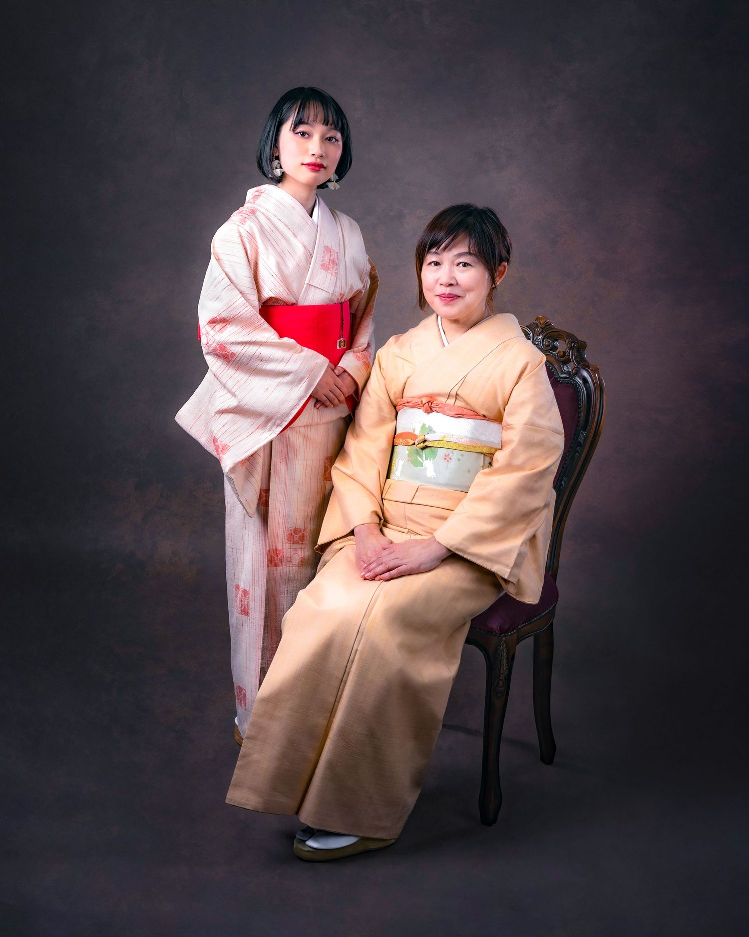Studio portrait of two people wearing traditional Japanese kimonos in pink and peach colors against a dark background.