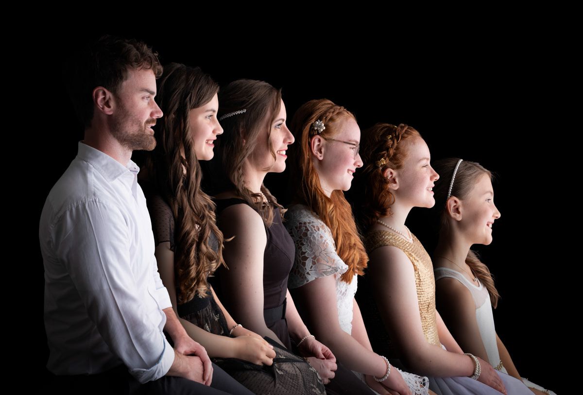 A line of performers in profile stand together against a black background, singing or performing in unison.