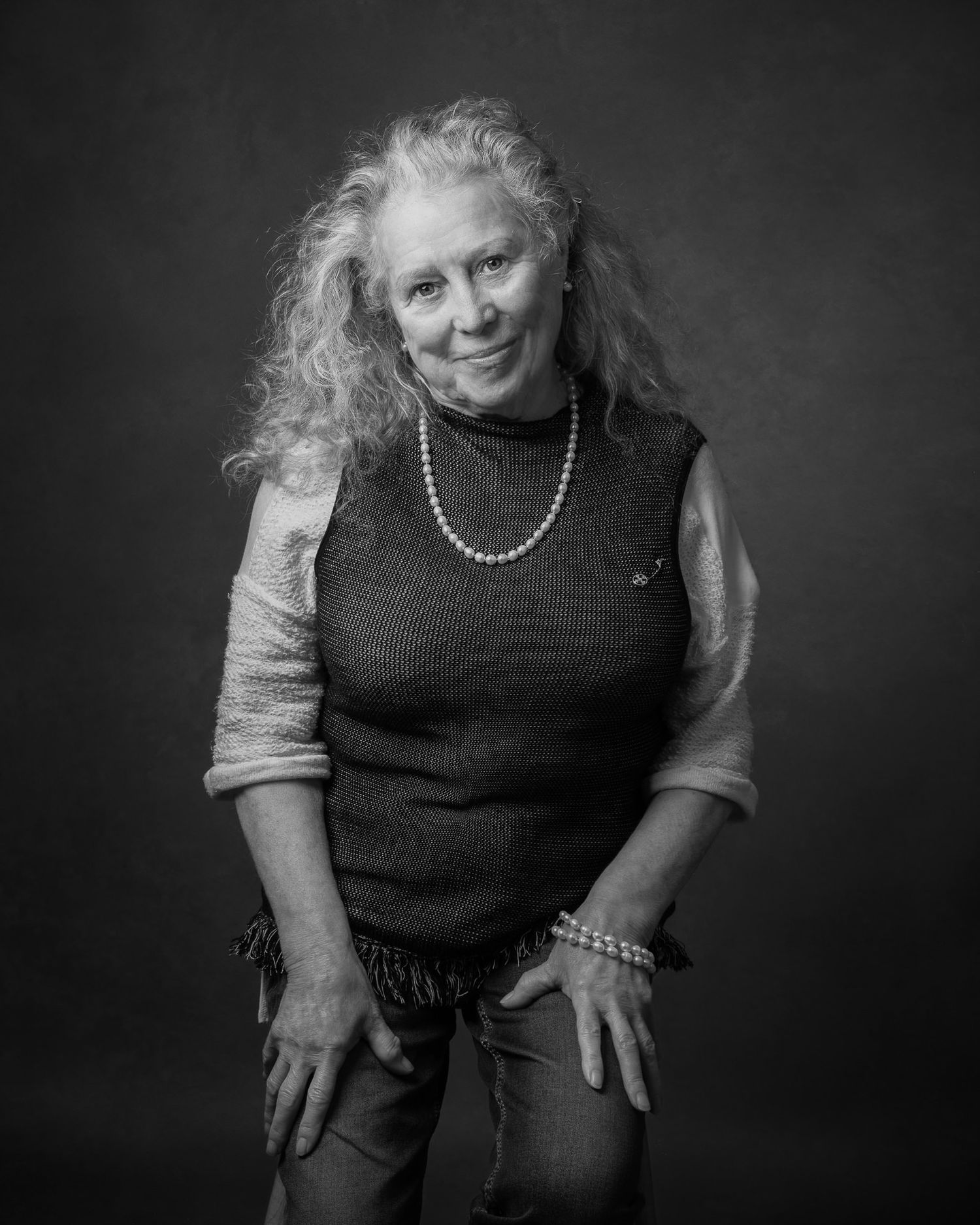 Black and white portrait of a person with curly hair wearing a dark sleeveless top and pearl necklace against dark background.
