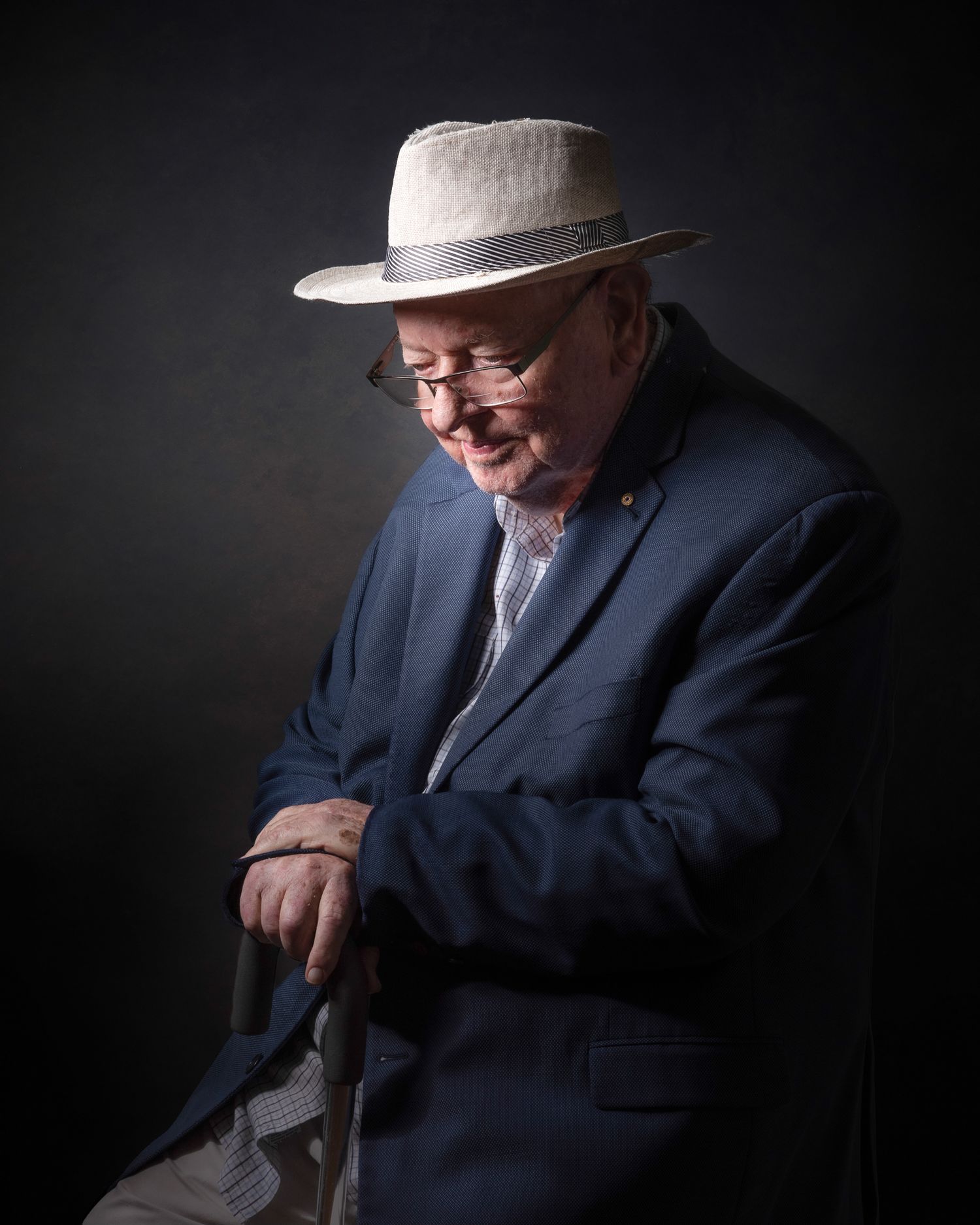 A portrait in moody lighting shows an elderly person in a grey fedora and navy blazer seated against a dark background.