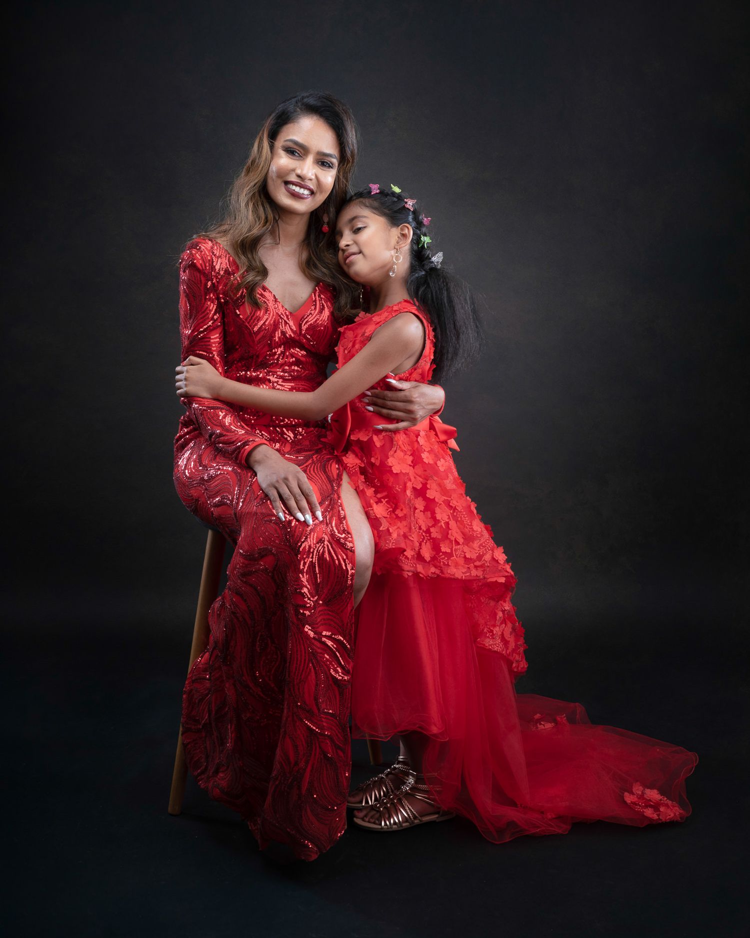 Two people in elegant red lace dresses pose together against a dark background for a formal portrait photo shoot.