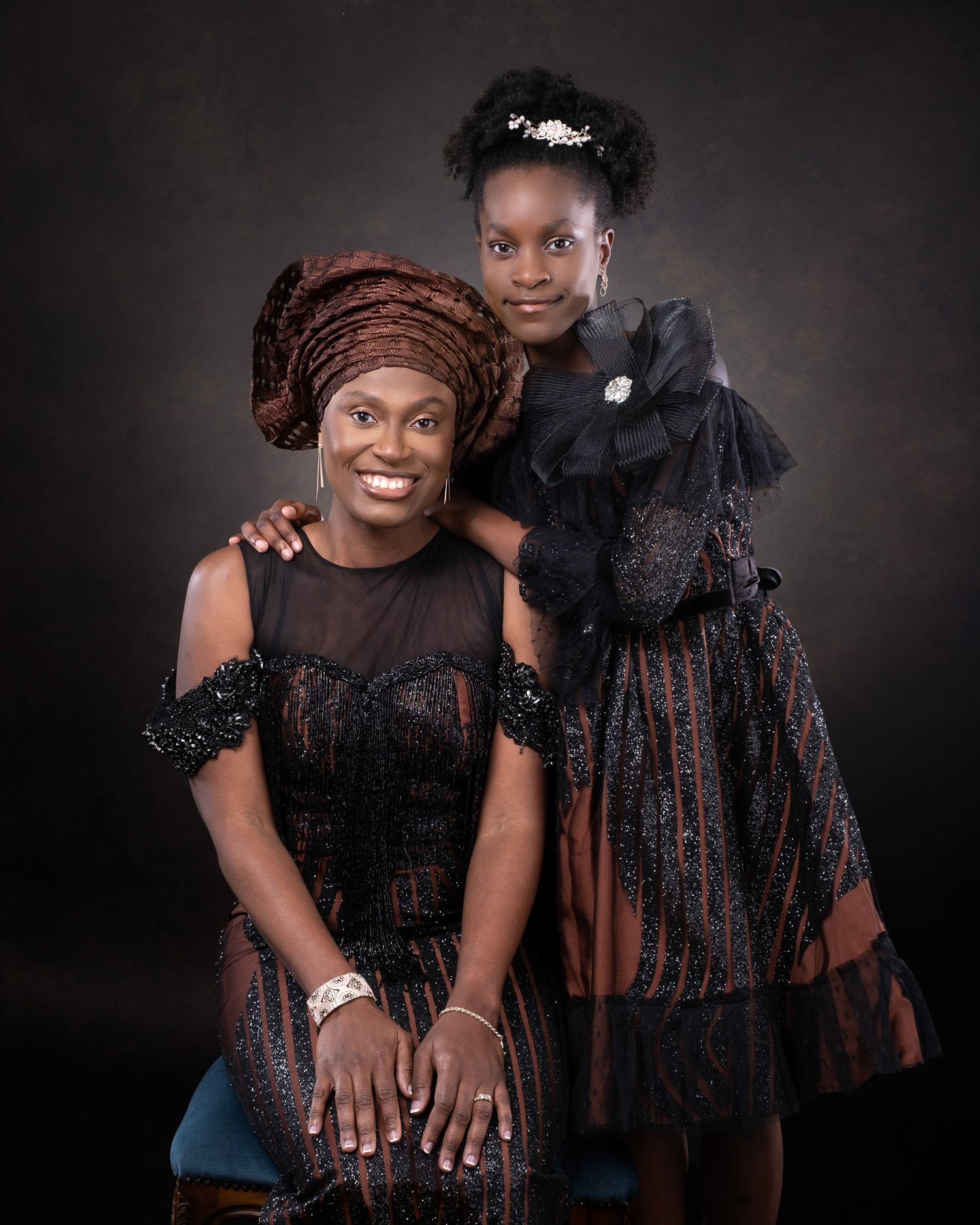 A portrait of two people wearing elegant black lace dresses against a dark background, one seated and one standing.