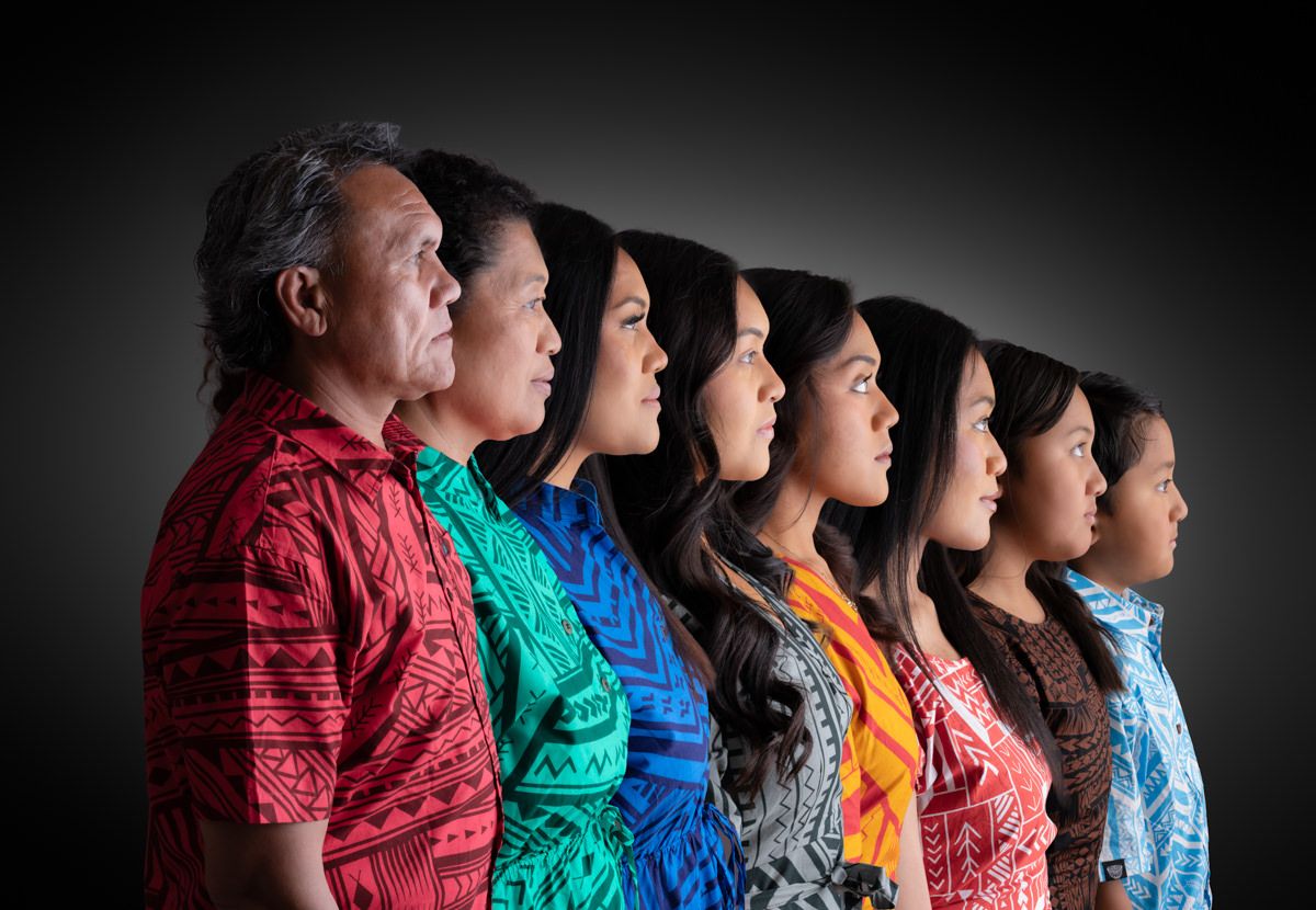 A group of people wearing colorful shirts stand in a line against a dark background for a dramatic portrait.
