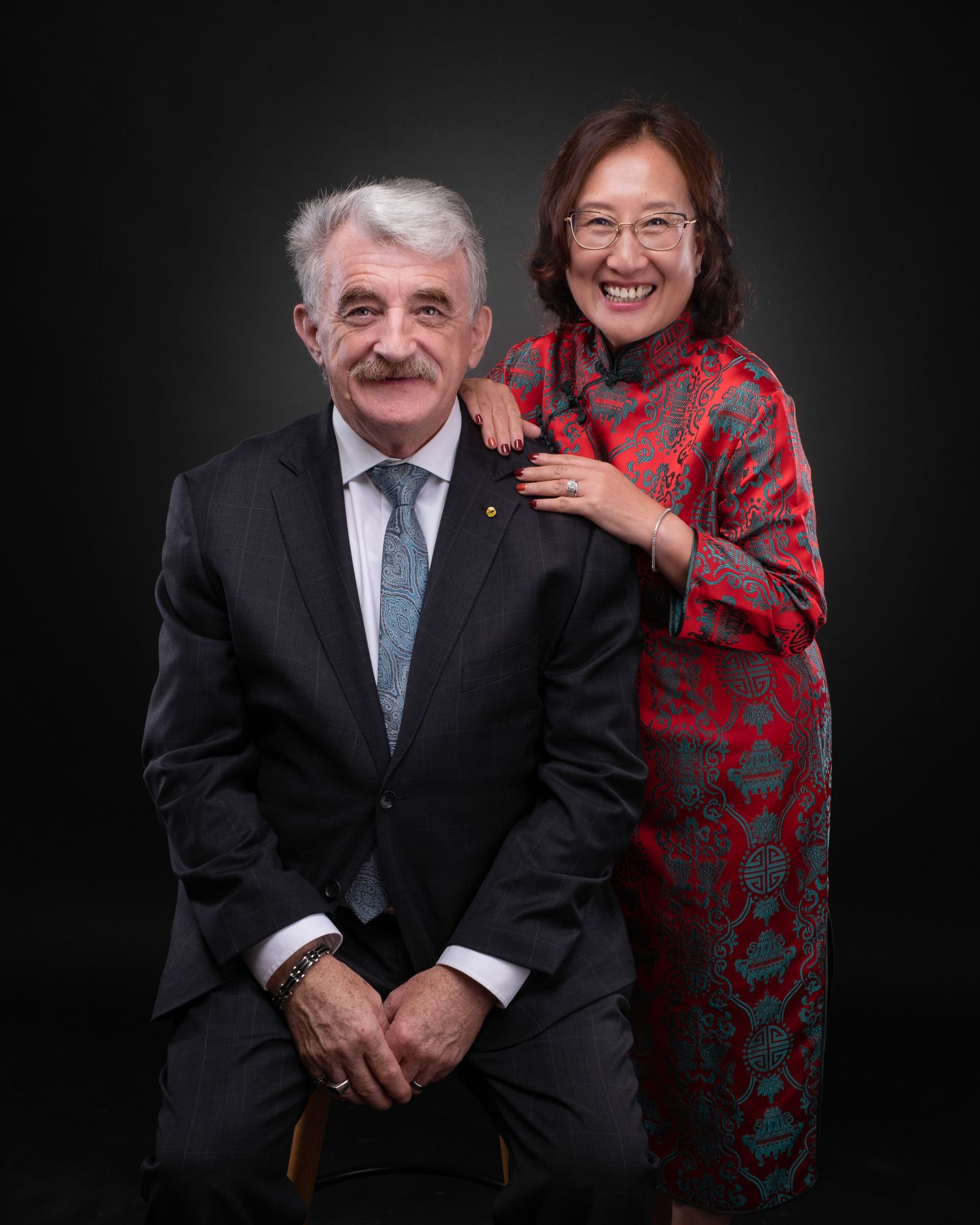 A professional portrait of two people posing against a dark background, one wearing a suit and tie and another in a red dress.