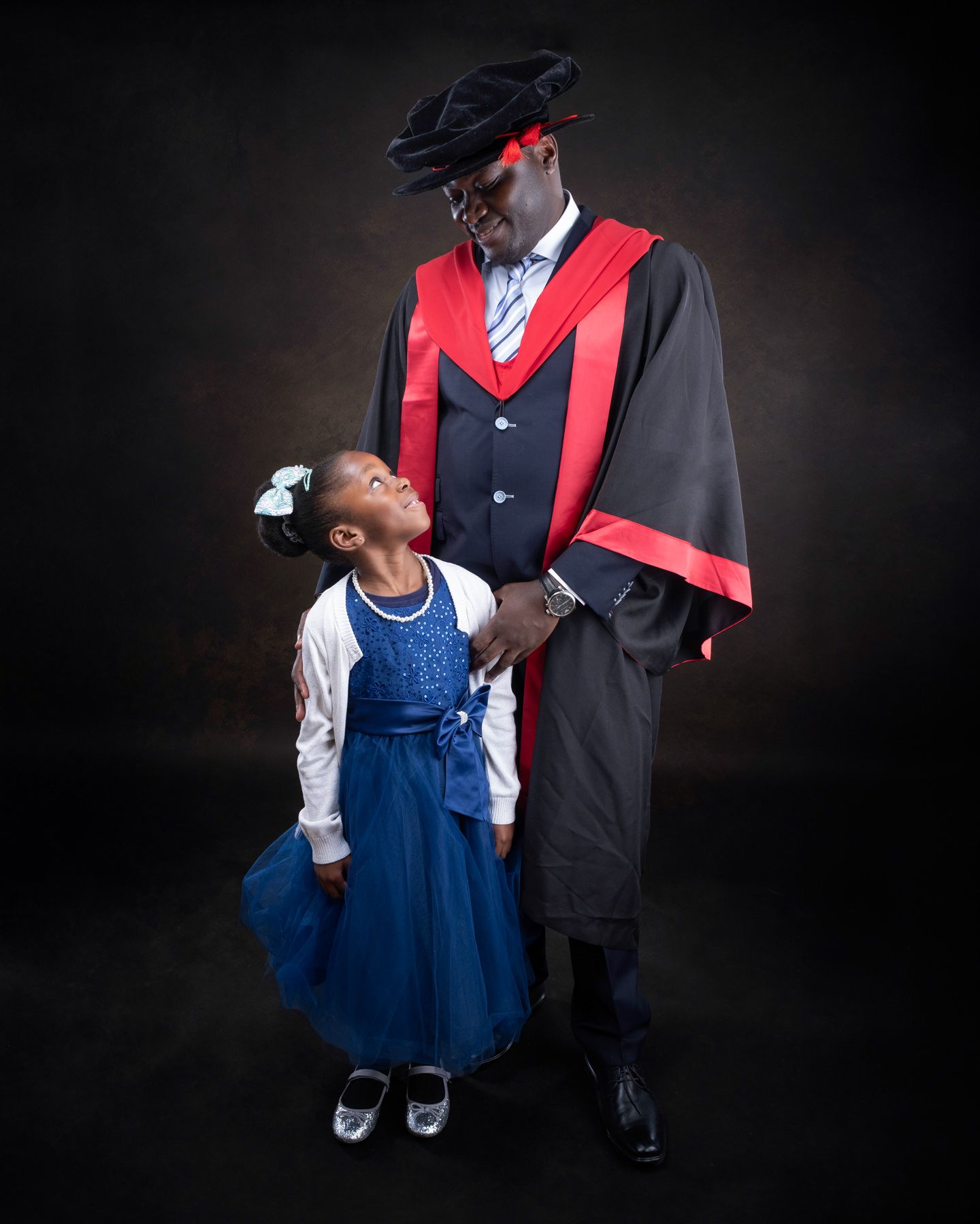 A couple in formal attire, with a blue dress and graduation gown, pose together against a dark background.