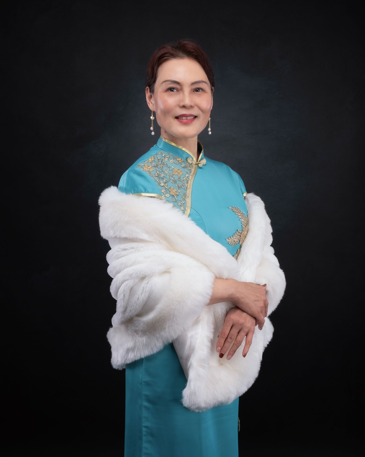 Person wearing a turquoise traditional Chinese qipao dress with gold embellishments and white fur wrap against dark background.