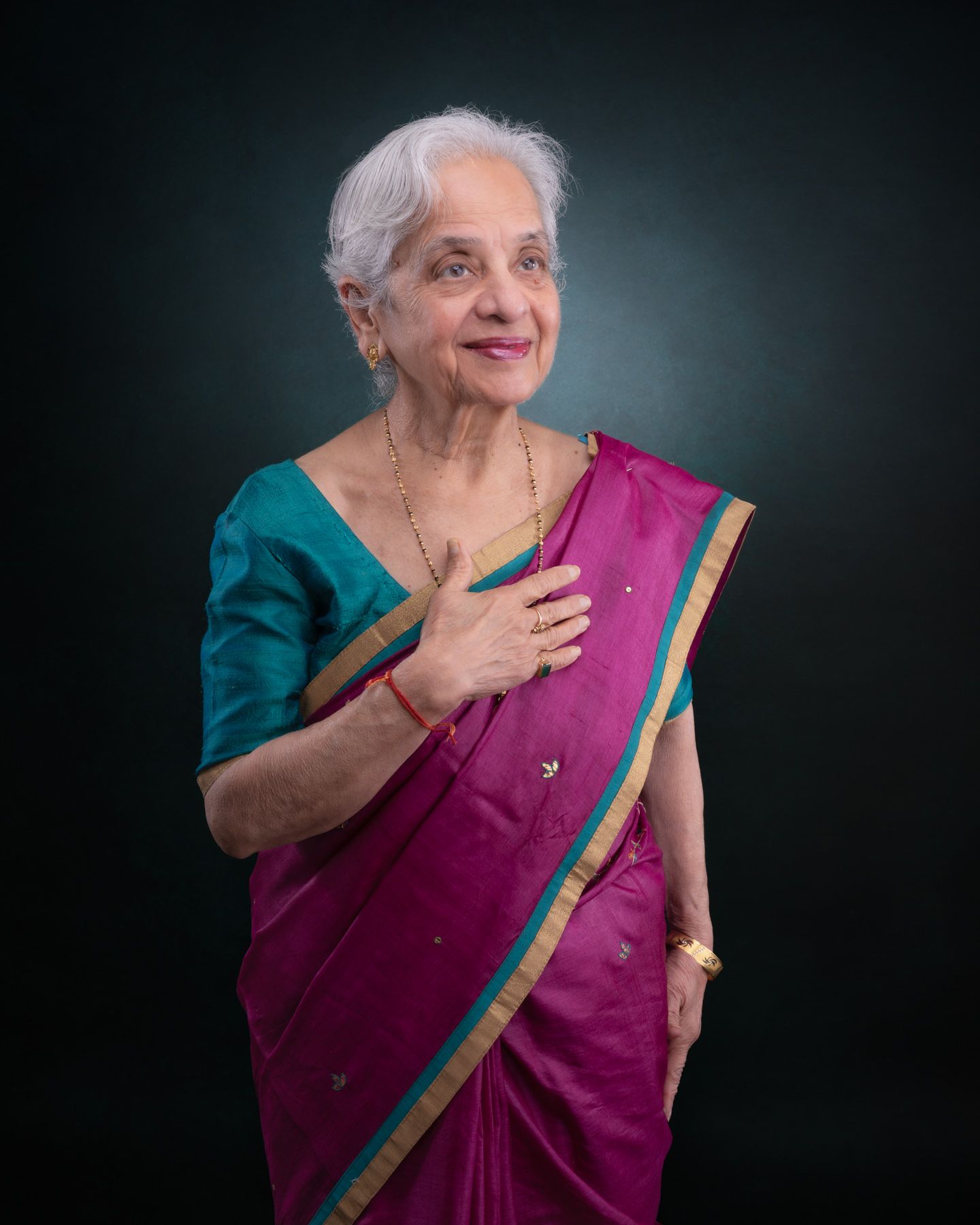 An elderly person in a purple sari with green trim poses expressively against a dark studio backdrop.