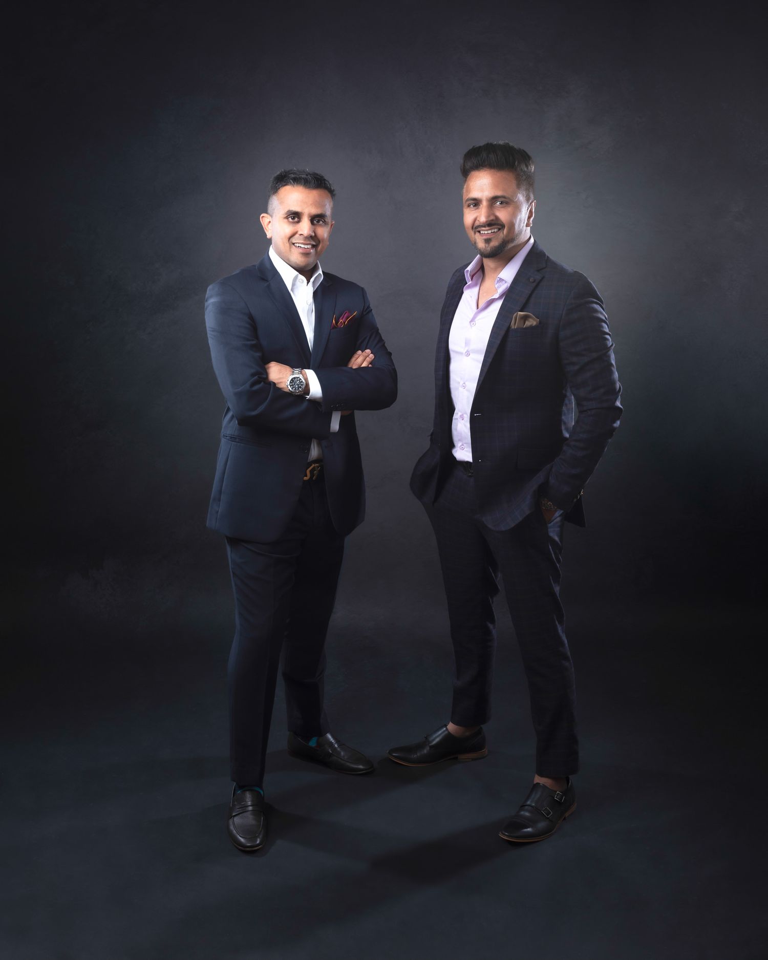 Two people in dark suits strike playful poses against a black background in a professional photo studio setting.
