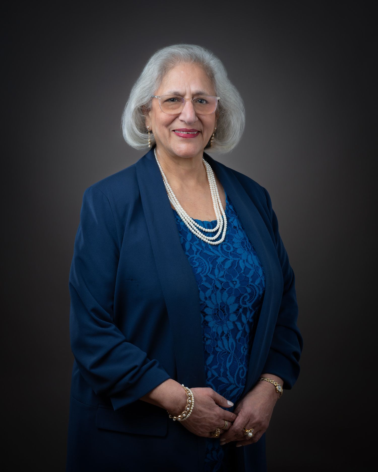 A senior woman wearing a navy blue blazer and pearl necklace poses elegantly against a dark backdrop.