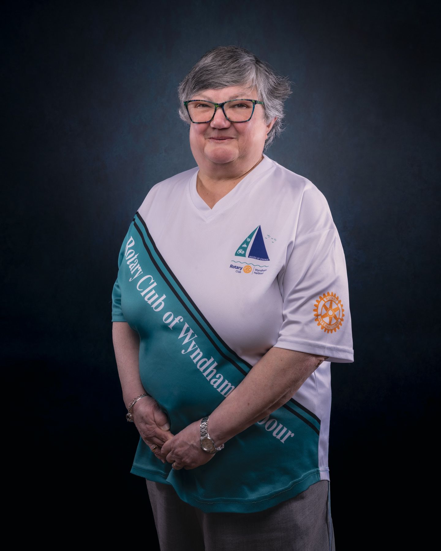 Studio portrait of someone wearing glasses and a white polo shirt with a teal sash against a dark background.