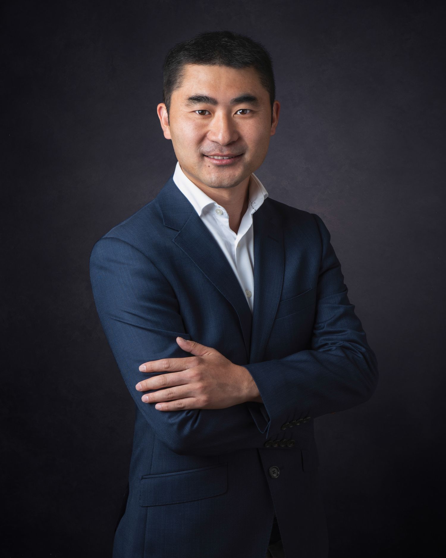 Professional studio portrait of someone in a navy suit jacket and white dress shirt against a dark background.