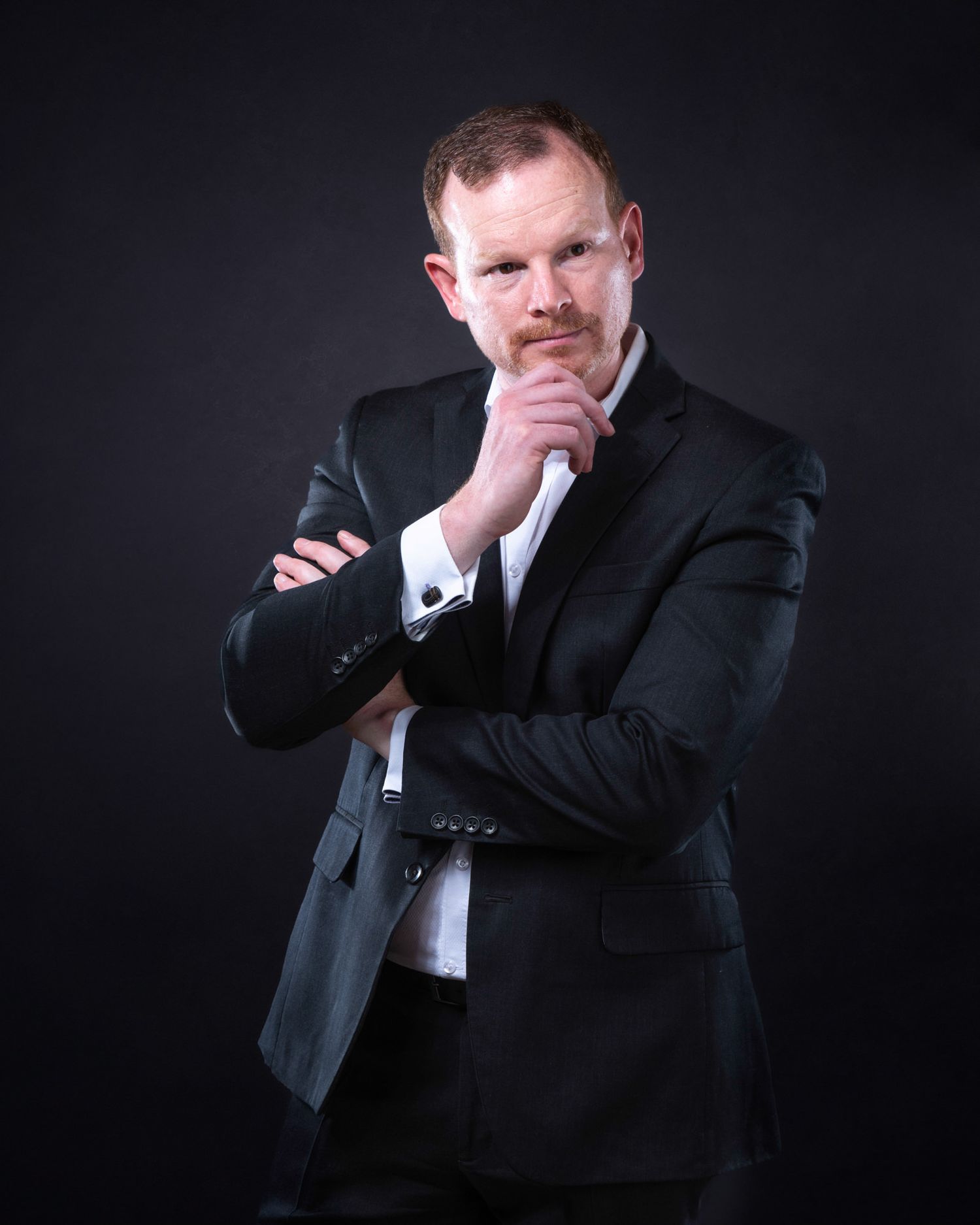 Professional studio portrait of someone in dark suit with thoughtful expression against black backdrop.