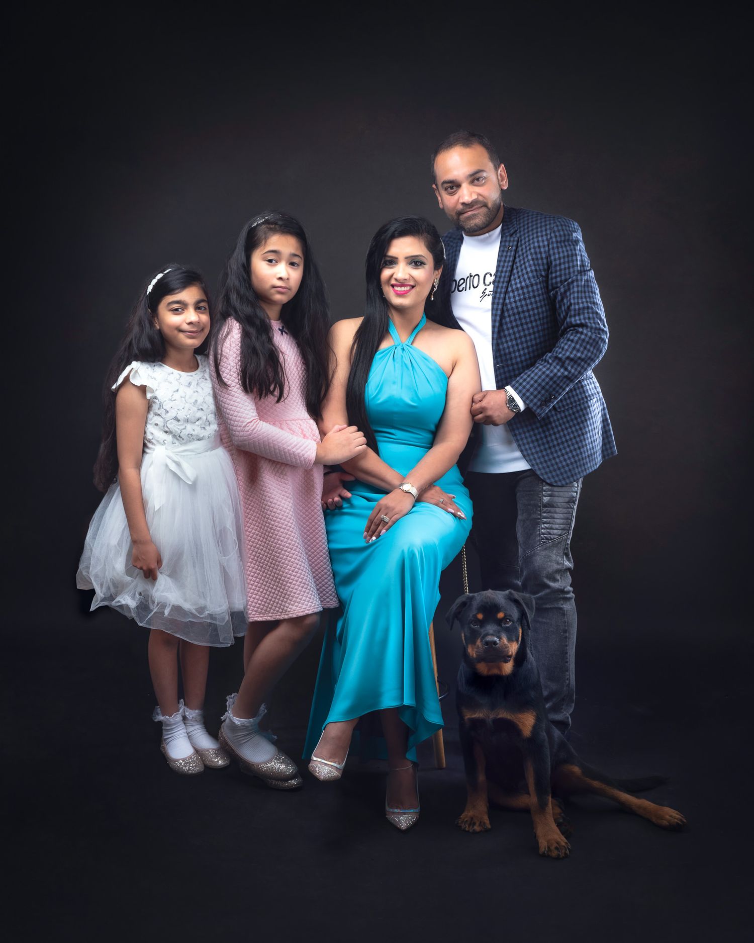 A stylish family in formal attire poses for a classic portrait against a dark background with their Rottweiler dog.