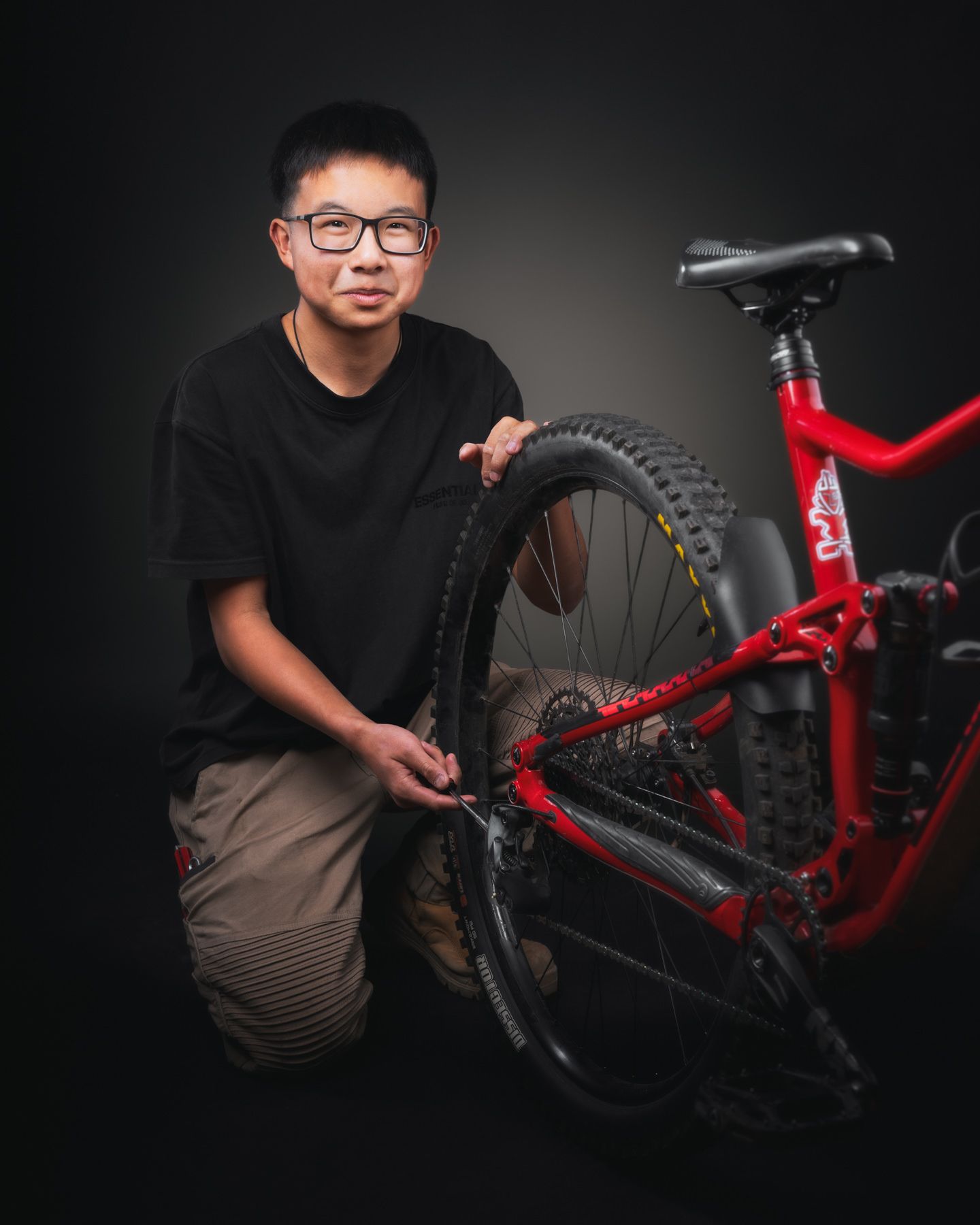 Portrait of someone kneeling next to a red mountain bike against a dark background in a studio setting.