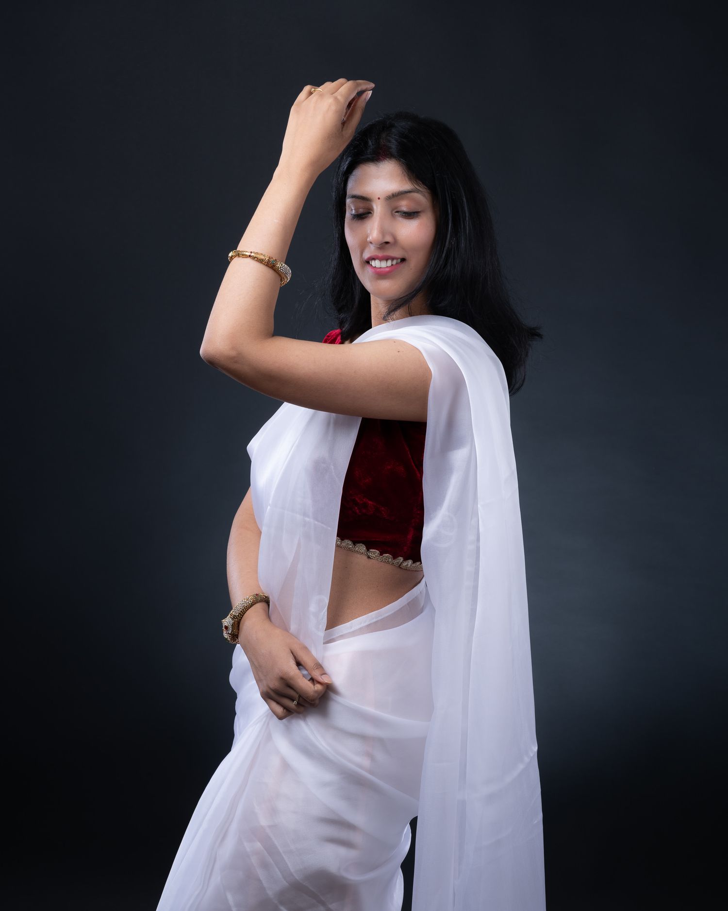 A woman poses gracefully in a white sari with a red blouse against a dark background in a studio setting.