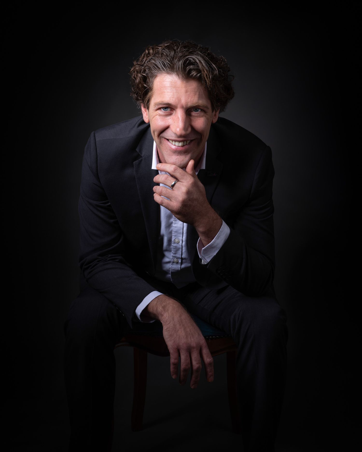 Professional studio portrait against dark background of someone in a dark suit sitting casually on a chair with a warm smile.